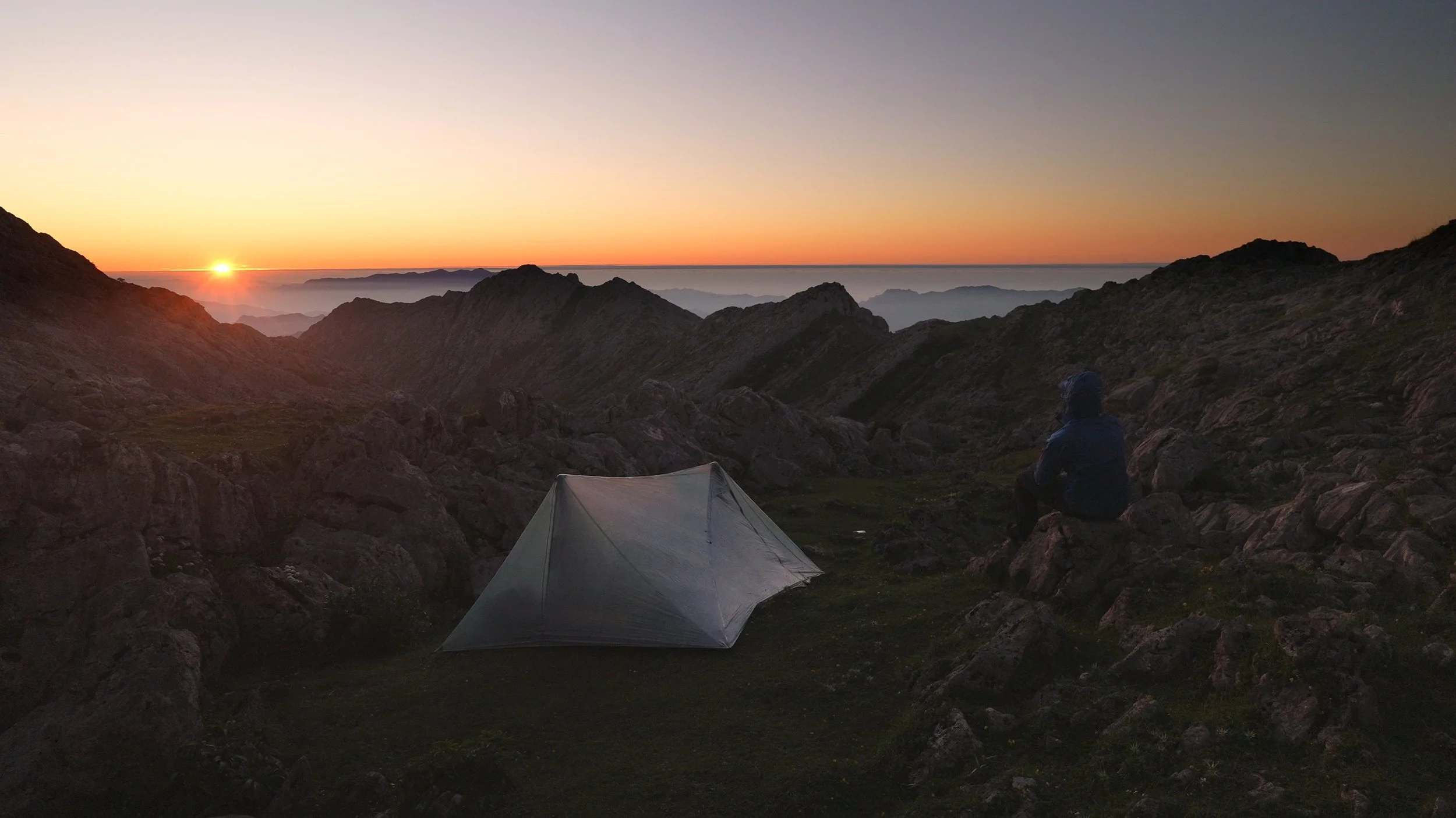 A person sitting on a rock in a mountainous area during sunrise, with a tent nearby and a landscape of rugged peaks and valleys.