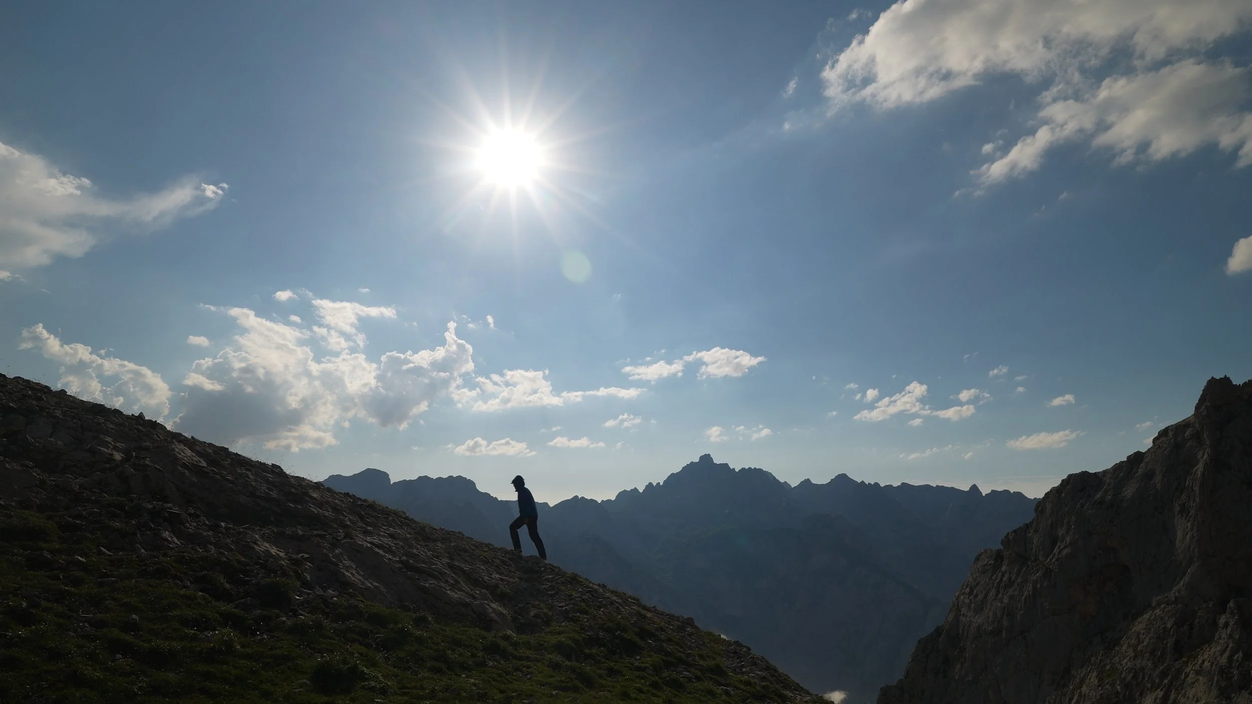 A person hiking on a mountain slope with a background of mountain peaks and a blue sky with clouds, bright sun shining.