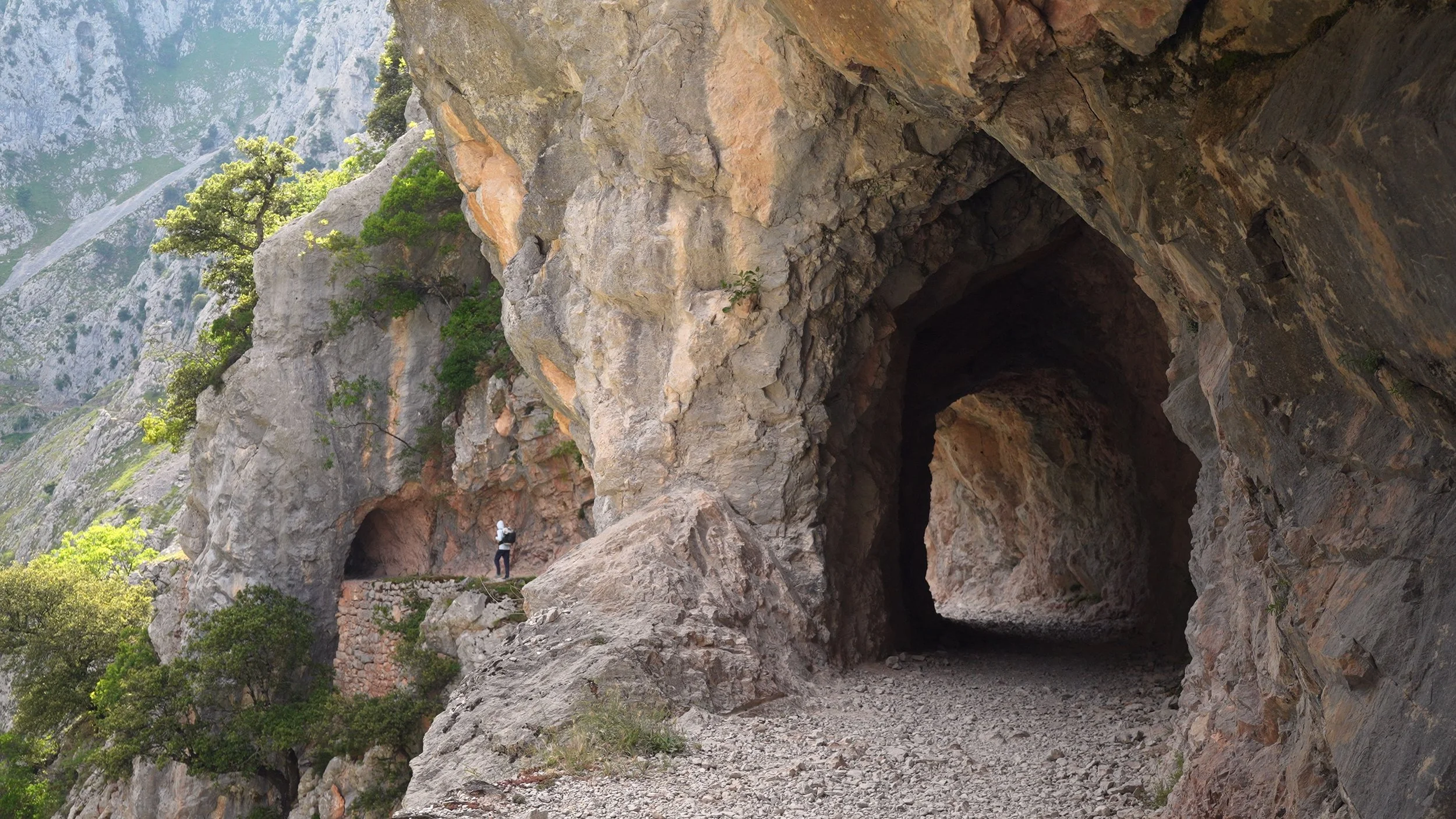 A person walking on a narrow mountain trail through a large rocky cave with lush green vegetation on the mountainside.