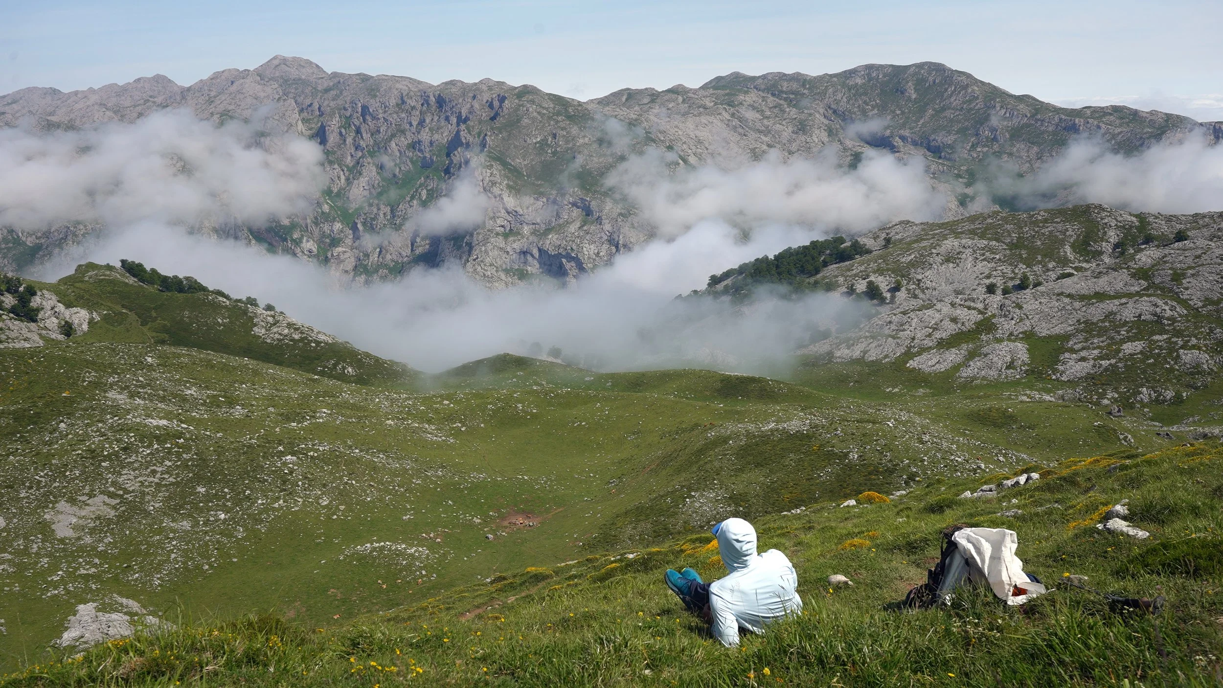 A person in a white hoodie sitting on grassy hillside overlooking green mountains with clouds and rocky peaks in the distance. A backpack is on the ground nearby.