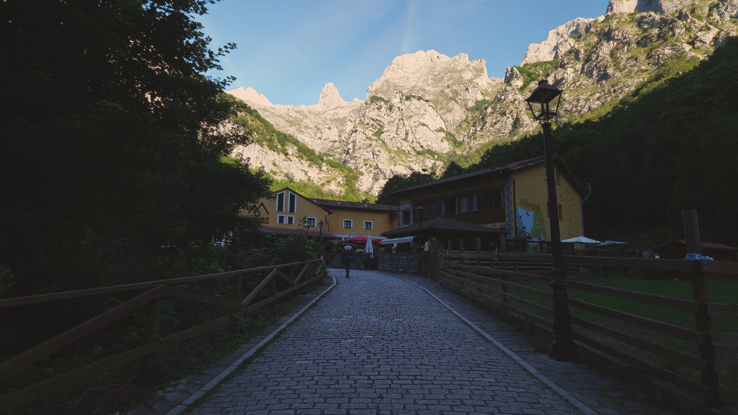A cobblestone path leading to a village in a mountainous area with rocky peaks and some greenery. There are buildings with signs and umbrellas along the path, and a person walking away from the camera.