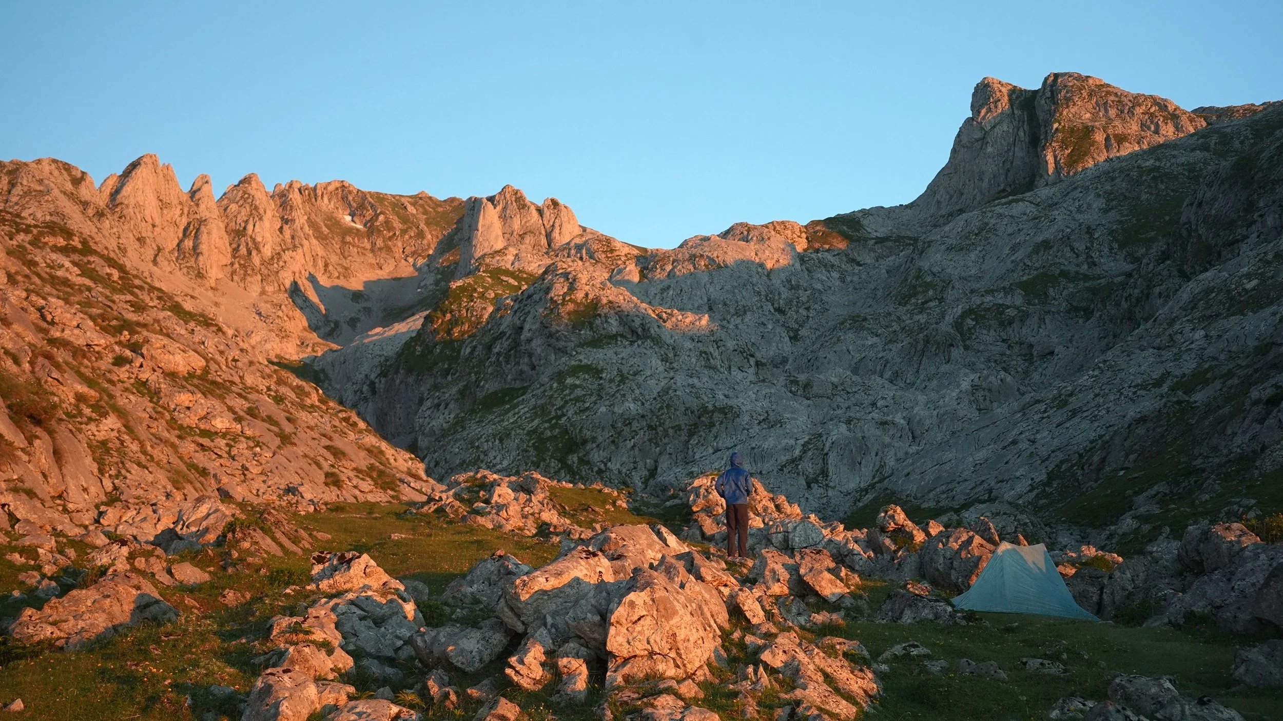 A person standing on rocks in a mountain landscape with rugged peaks, a small tent, and a clear blue sky