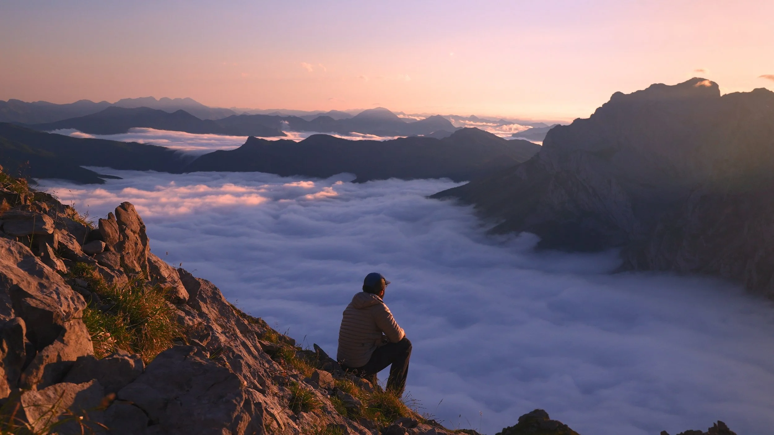 A person sitting on a rocky mountain slope watching a sunrise over a sea of clouds and mountain ranges.