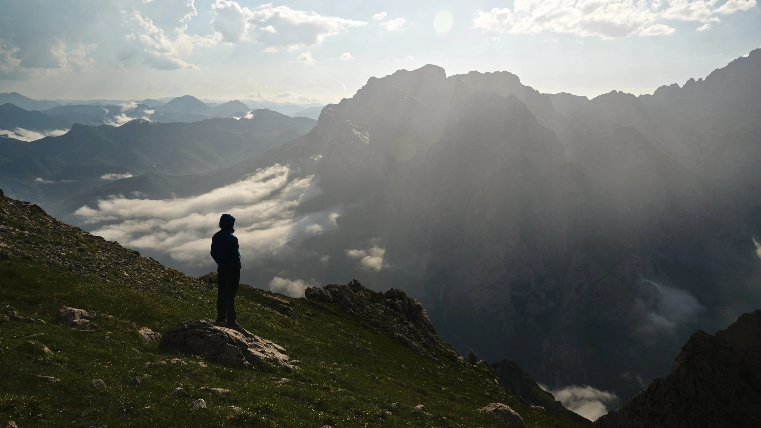 A person standing on a grassy slope with rocks, overlooking a mountain range with clouds and sunlight in the background.
