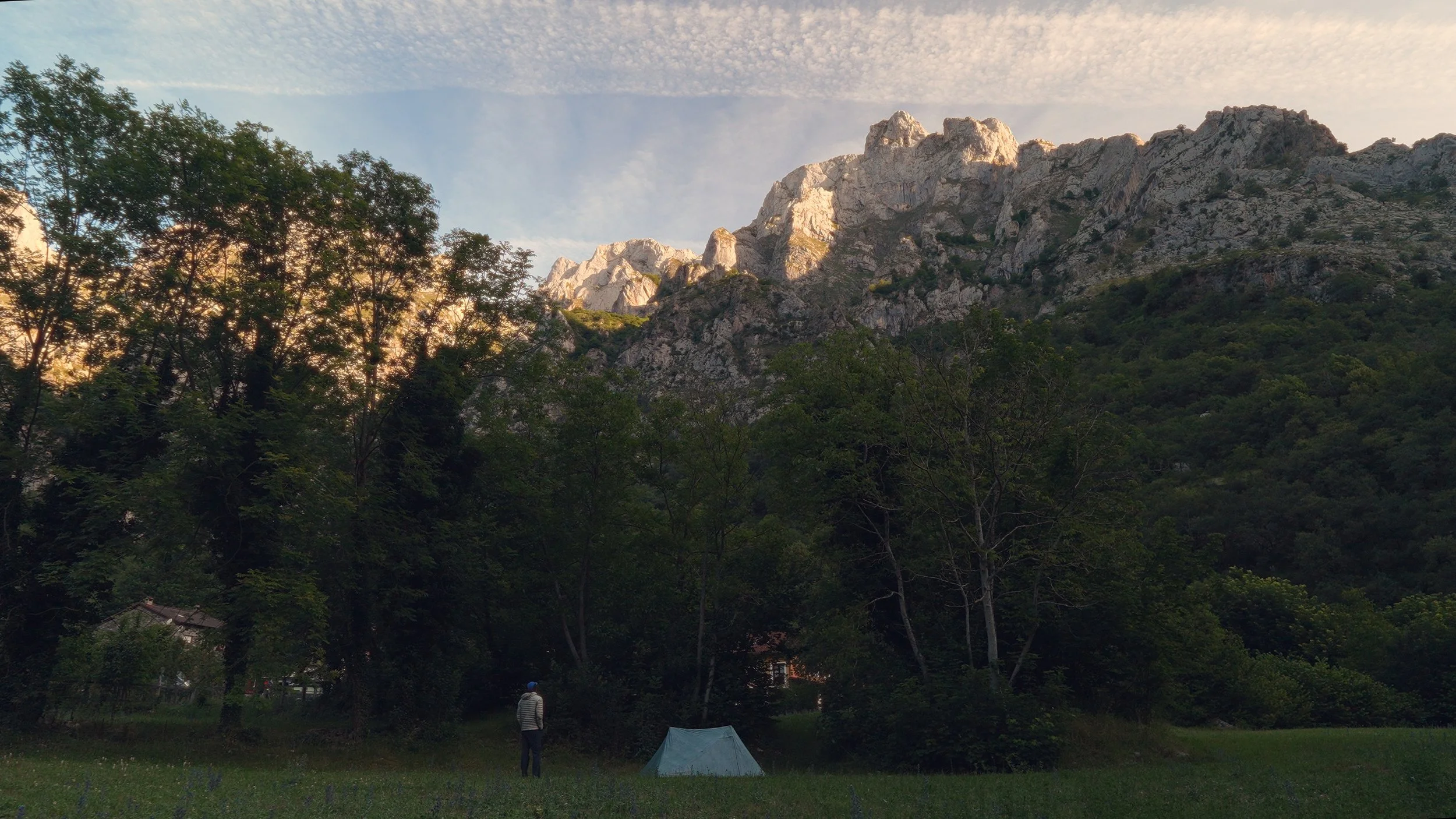 A man with a backpack standing near a tent in a grassy field, with lush trees and rocky mountains in the background under a partly cloudy sky at sunset.