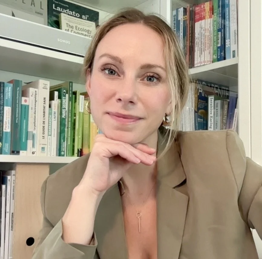A woman with long blond hair and fair skin, wearing a black blazer over a white blouse, stands in front of a bookshelf filled with colorful books and artwork, smiling softly.