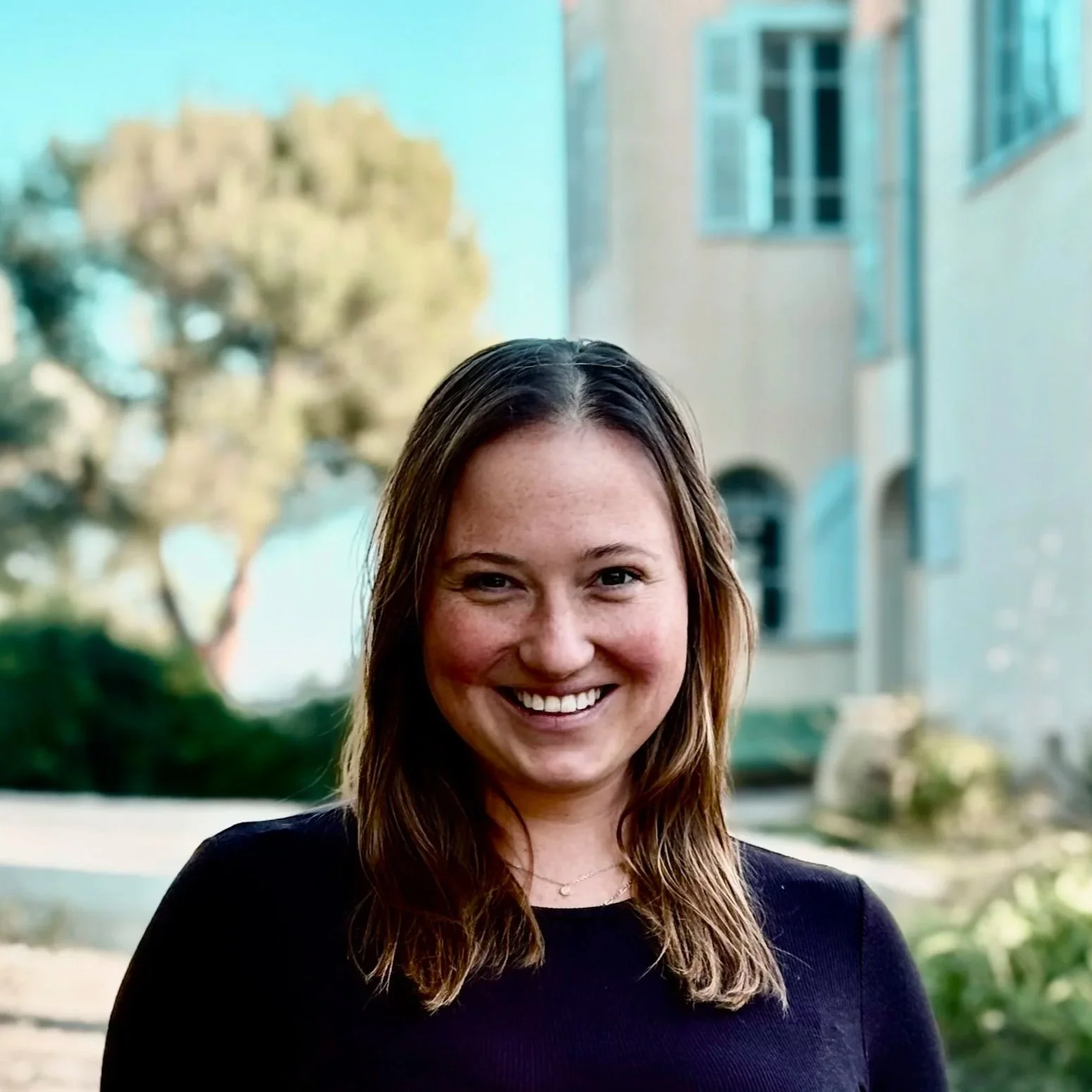 A smiling woman with shoulder-length brown hair standing outdoors in front of a blurred background of trees and a building.