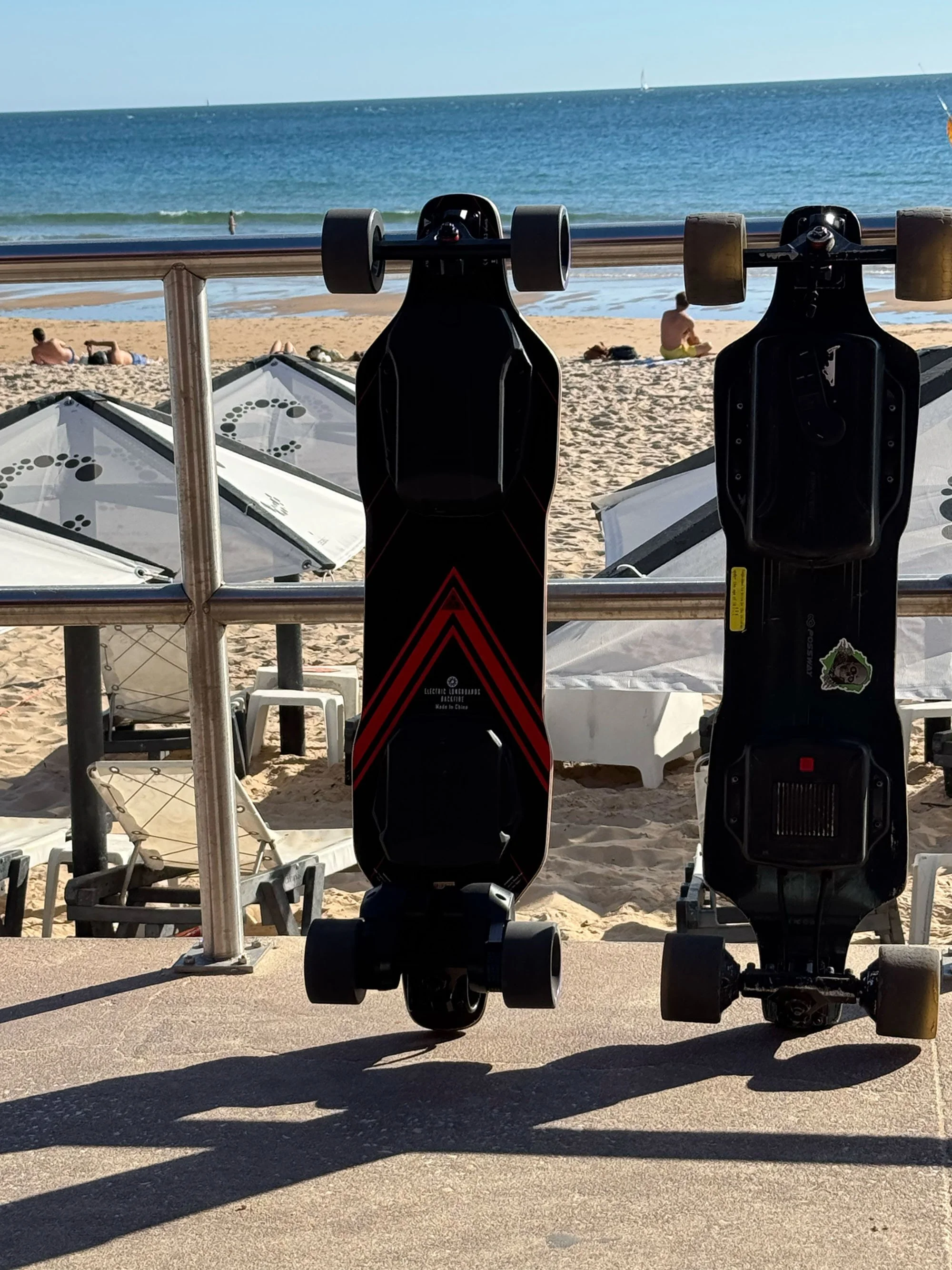 Two electric skateboards are mounted on a railing overlooking a beach with sand, a few people sitting and lying on the sand, and the ocean with sailboats in the distance.