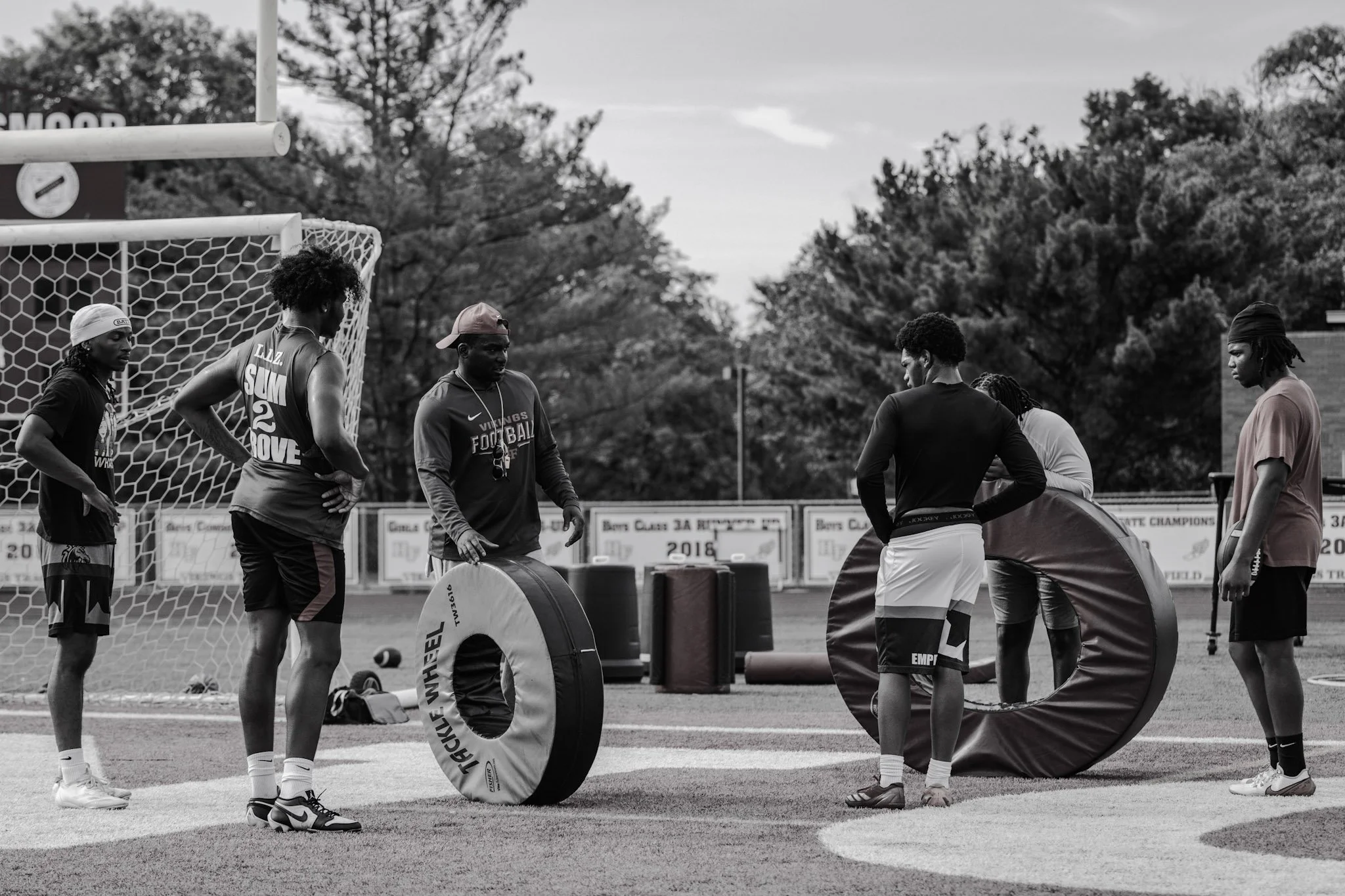 Group of young men on a football field preparing for practice, engaging in a drill with padded equipment.