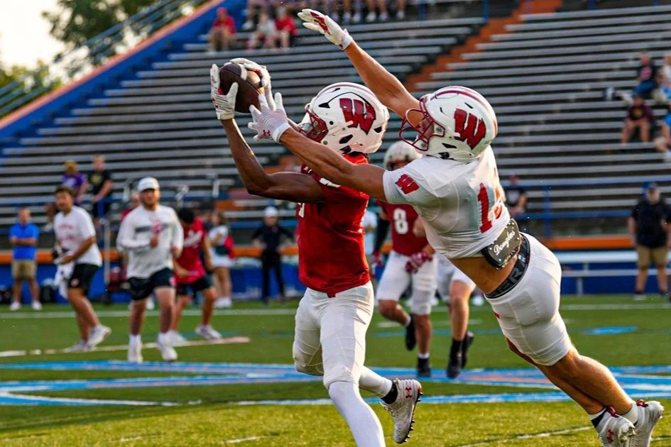 Two football players in white and red uniforms are competing for possession of the football during a game, with one player jumping to catch it while the other tries to block or intercept.