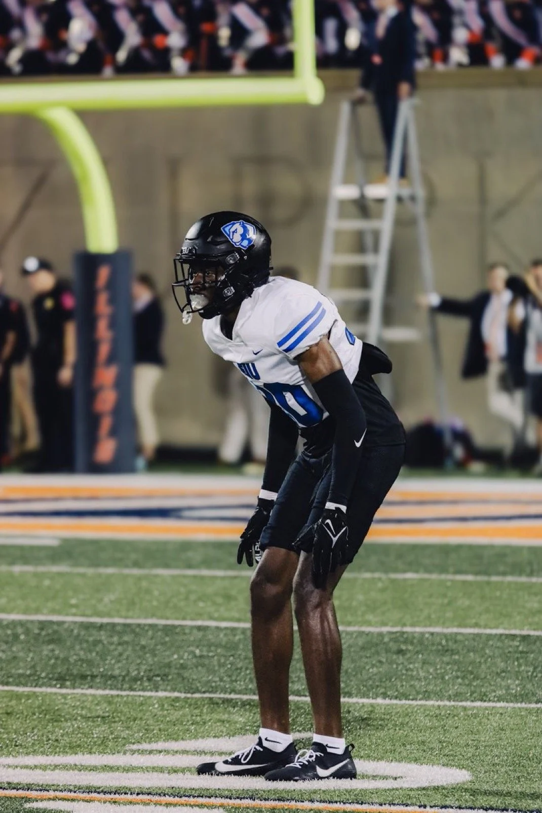 American football player in a white and blue uniform standing on the field, preparing for the next play, with a defensive stance.