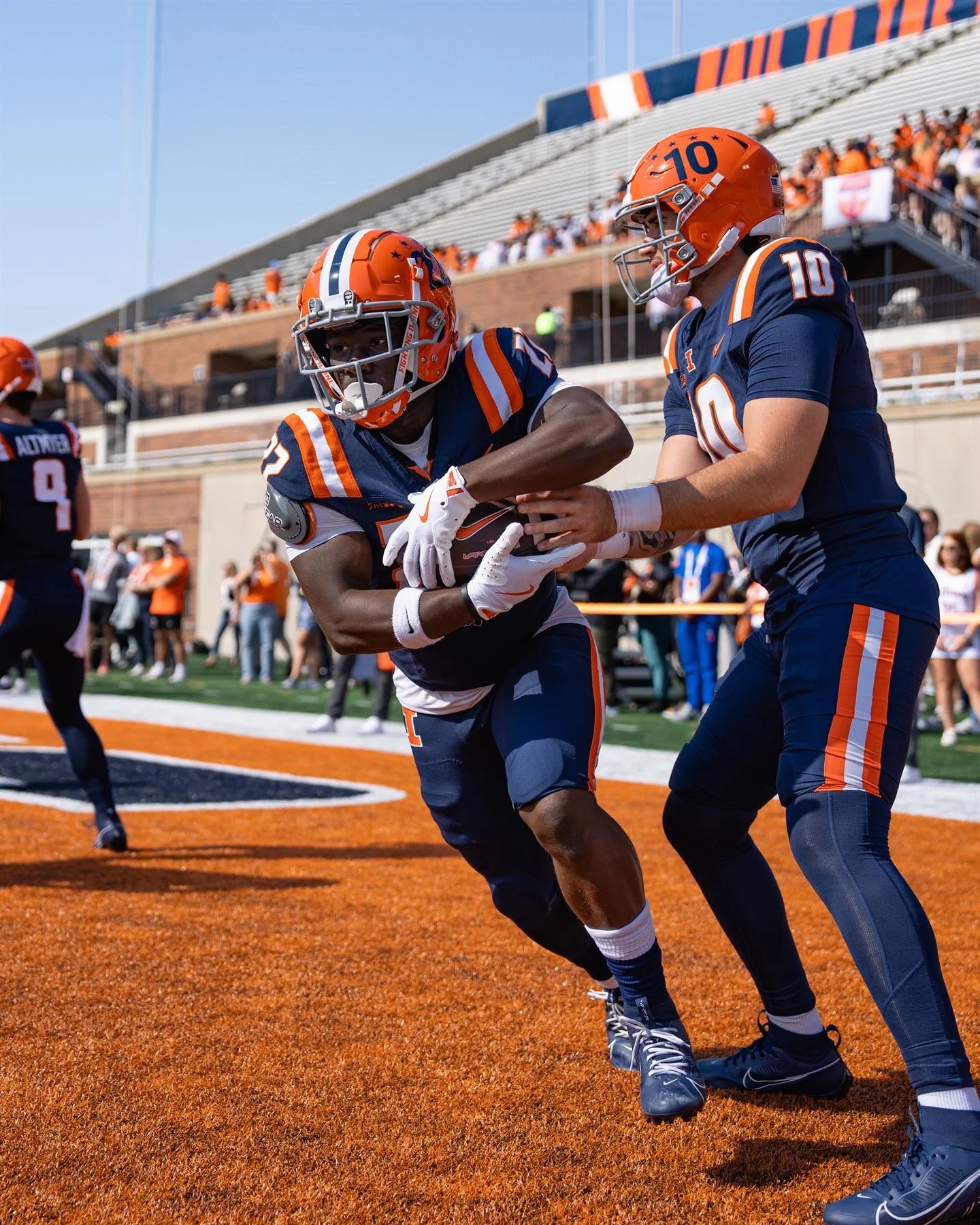 Two football players in orange and navy uniforms practicing on the field during a game, with one player holding the ball and a crowd watching in the background.