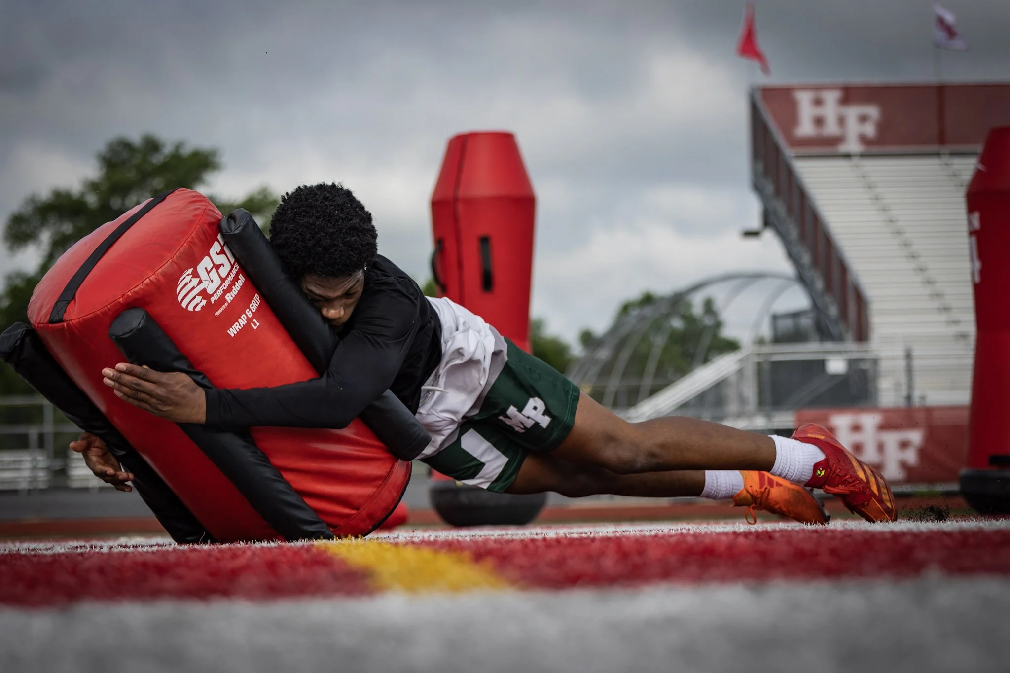 Football player practicing tackling drill on a red padded shield on an outdoor field with a cloudy sky in the background.