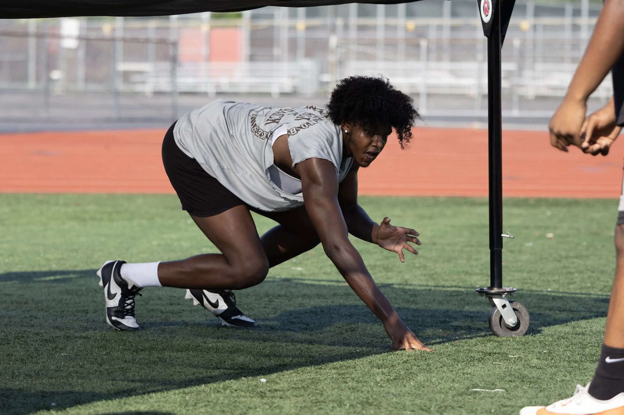 A female athlete is on the ground in a crawling position on a running track, with one hand on the grass and the other reaching forward, wearing athletic clothing and shoes.