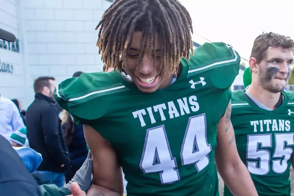Young football player with dreadlocks smiling, wearing a green jersey with the word 'TITANS' and the number 44, beside teammate in matching uniform.