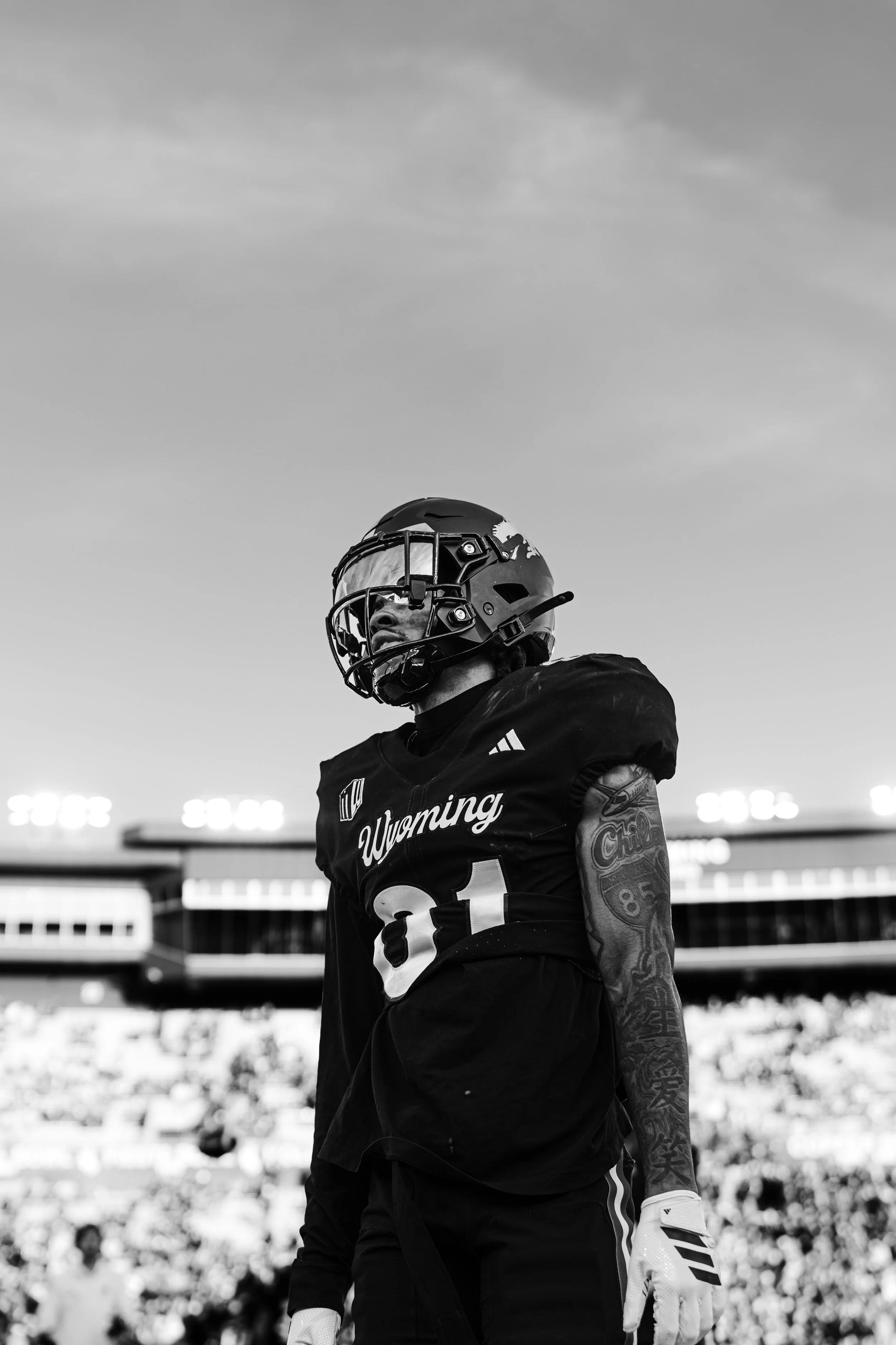 Black and white photo of a football player in uniform with a helmet and gloves, standing on a football field with a stadium and crowd in the background.
