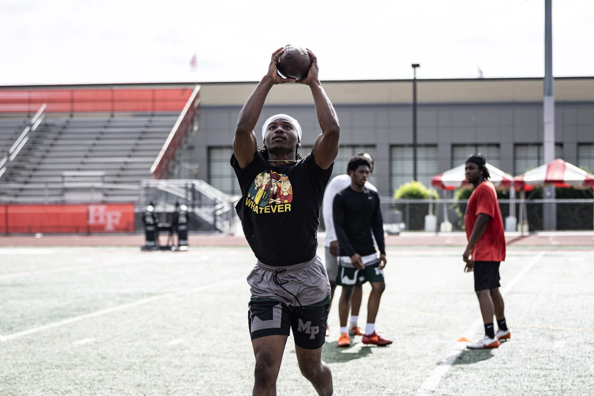 A young man catching a football during an outdoor practice, with three other men in the background on a sports field.