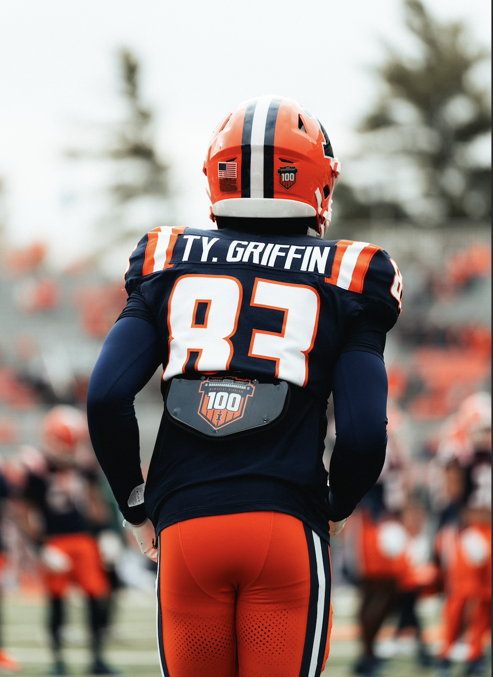 Back of American football player Ty Griffin wearing a navy jersey with orange and white accents, red pants, and a helmet with a Patriot logo, standing on the field during a game or practice.