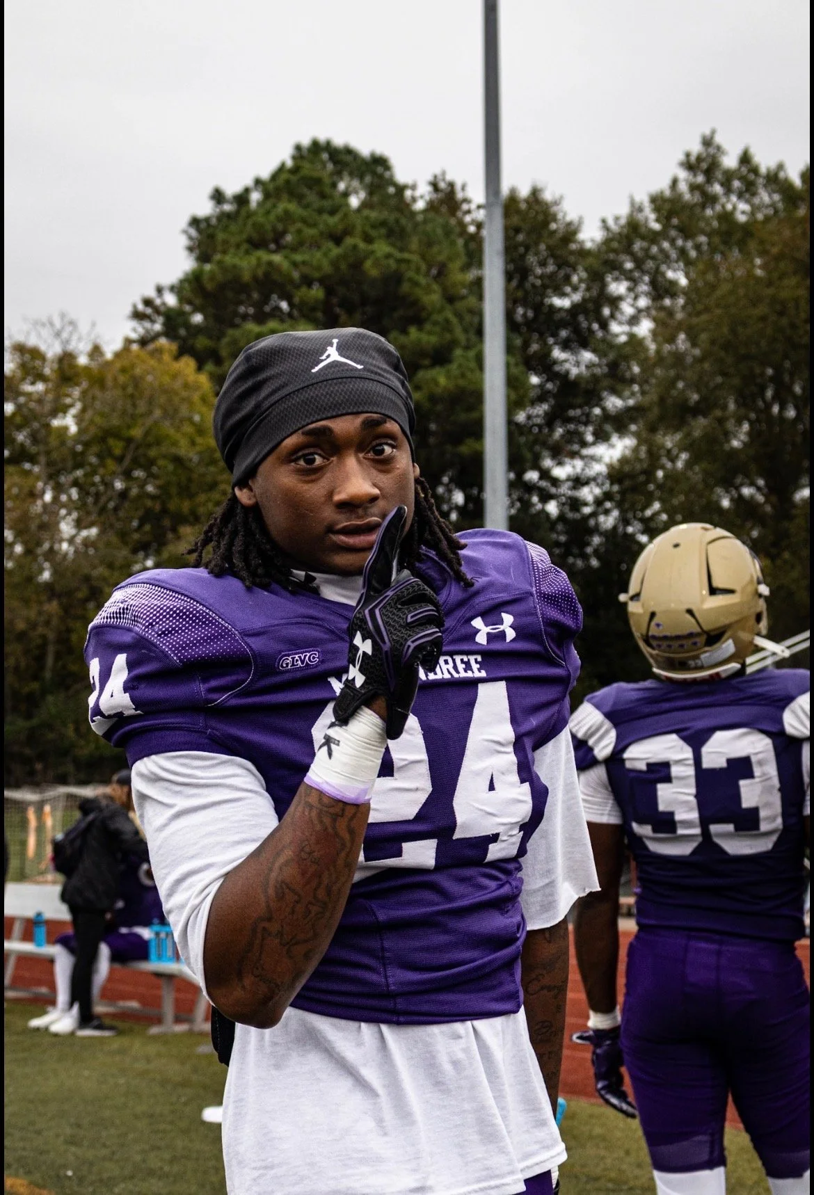 An African American football player with jersey number 44, wearing purple and white uniform, black gloves, and a black headband, is holding a finger to his lips in a shushing gesture on a football field during daytime.