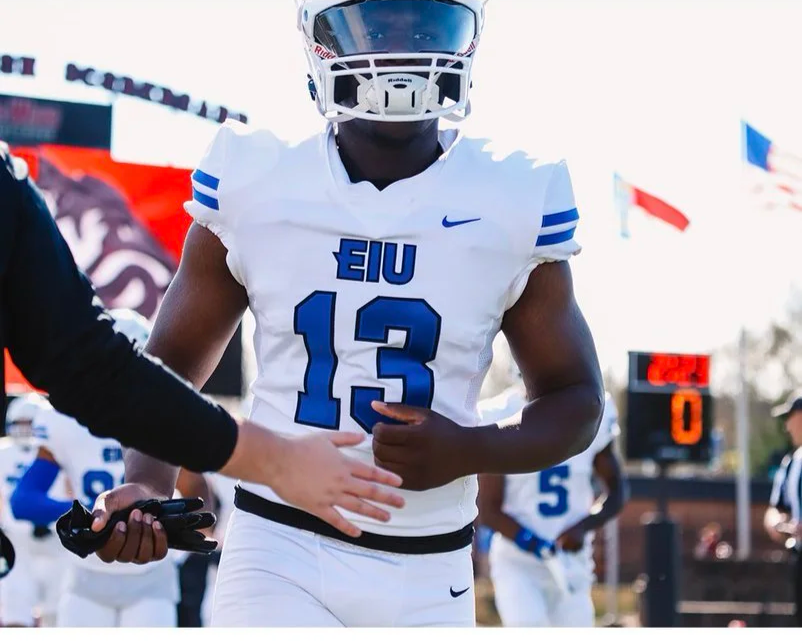 American football player wearing a white jersey with the number 13 and 'EIU' logo, holding a football, on the sidelines of a game with flags in the background.