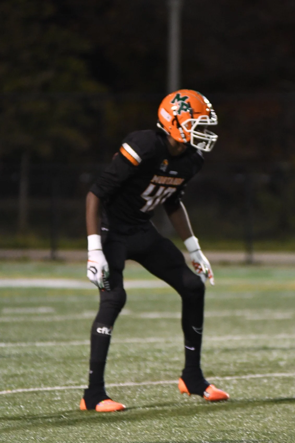 A football player wearing a black uniform and orange helmet standing on a football field at night.