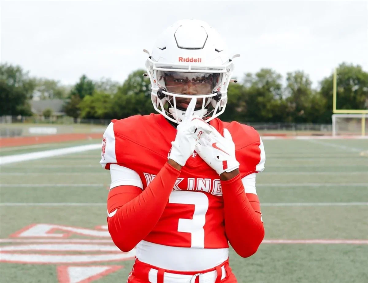 A football player in a red and white uniform, and helmet, standing on a football field making a shushing gesture.
