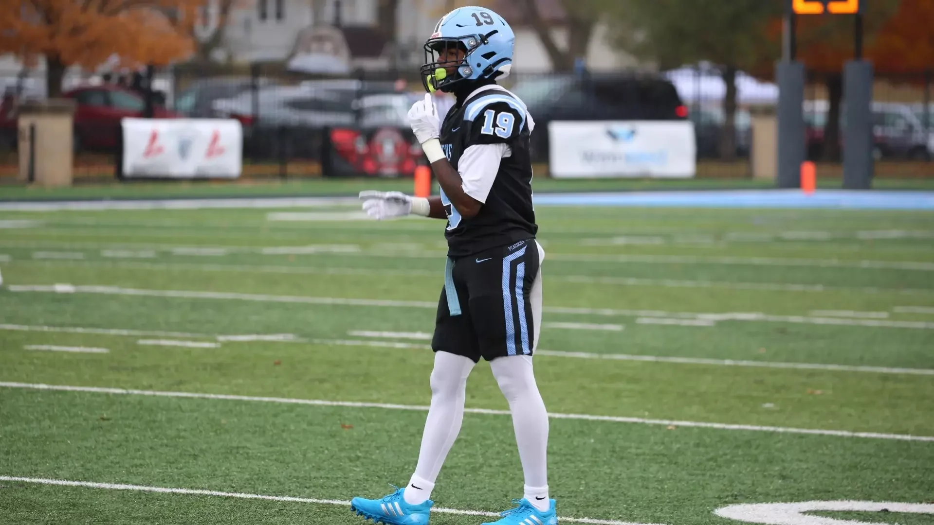 Football player standing on a field wearing a black and blue uniform with the number 19, a blue helmet, and light blue cleats, holding a yellow whistle near his mouth.