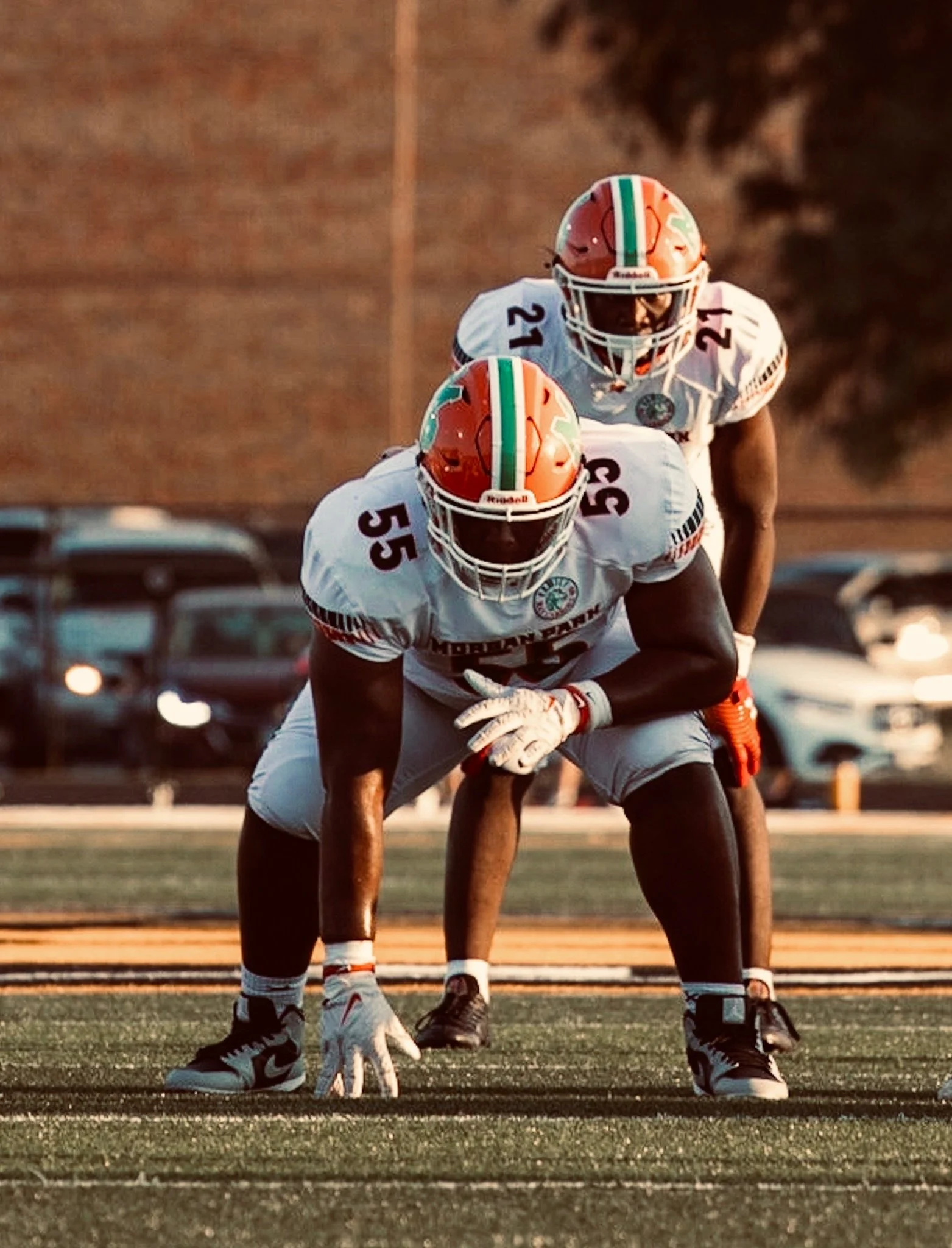 Two football players in white jerseys and orange helmets set for a snap on the field during sunset.