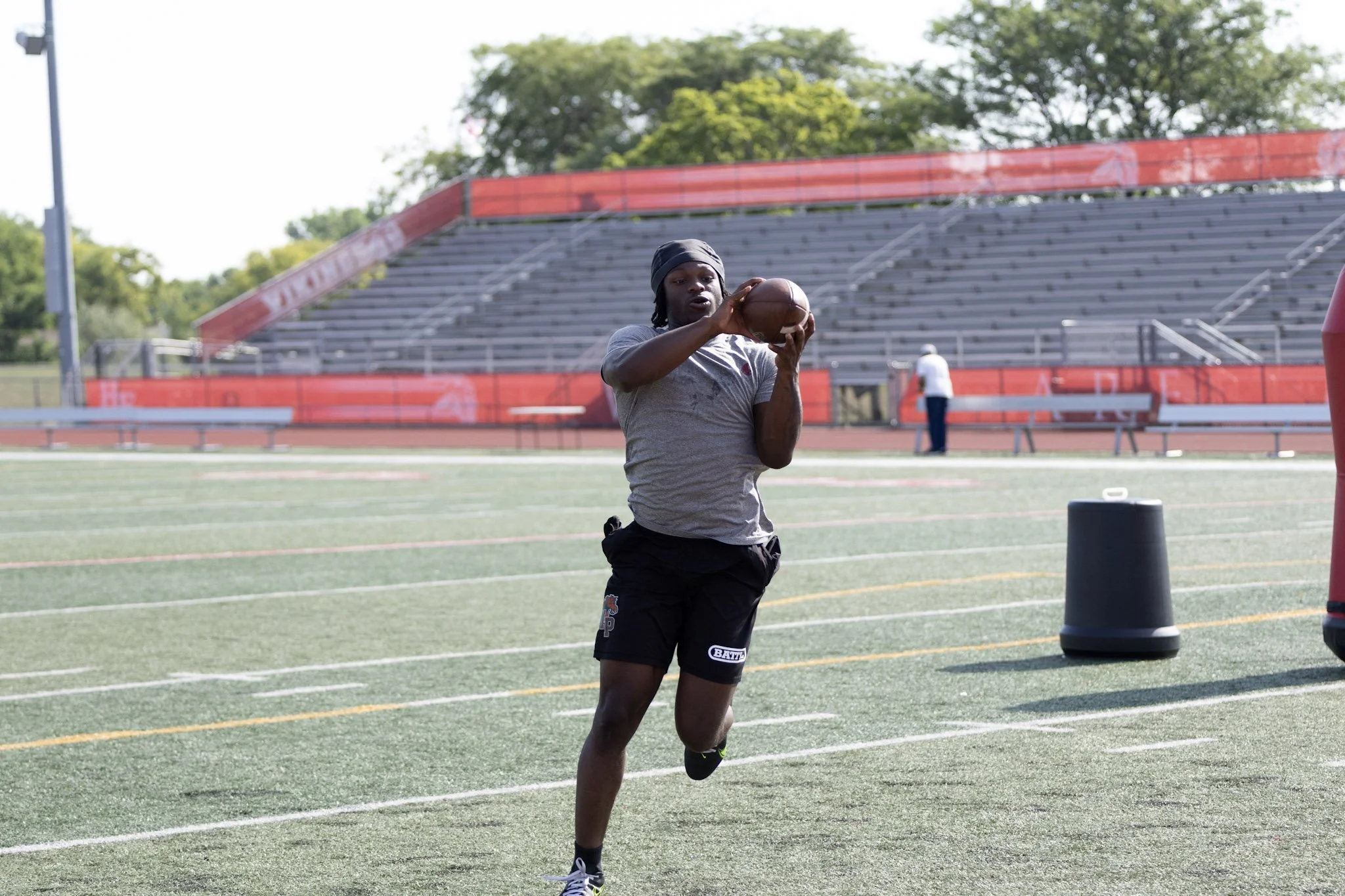 A man holding a football on a football field during practice, with empty bleachers and a person in the background.