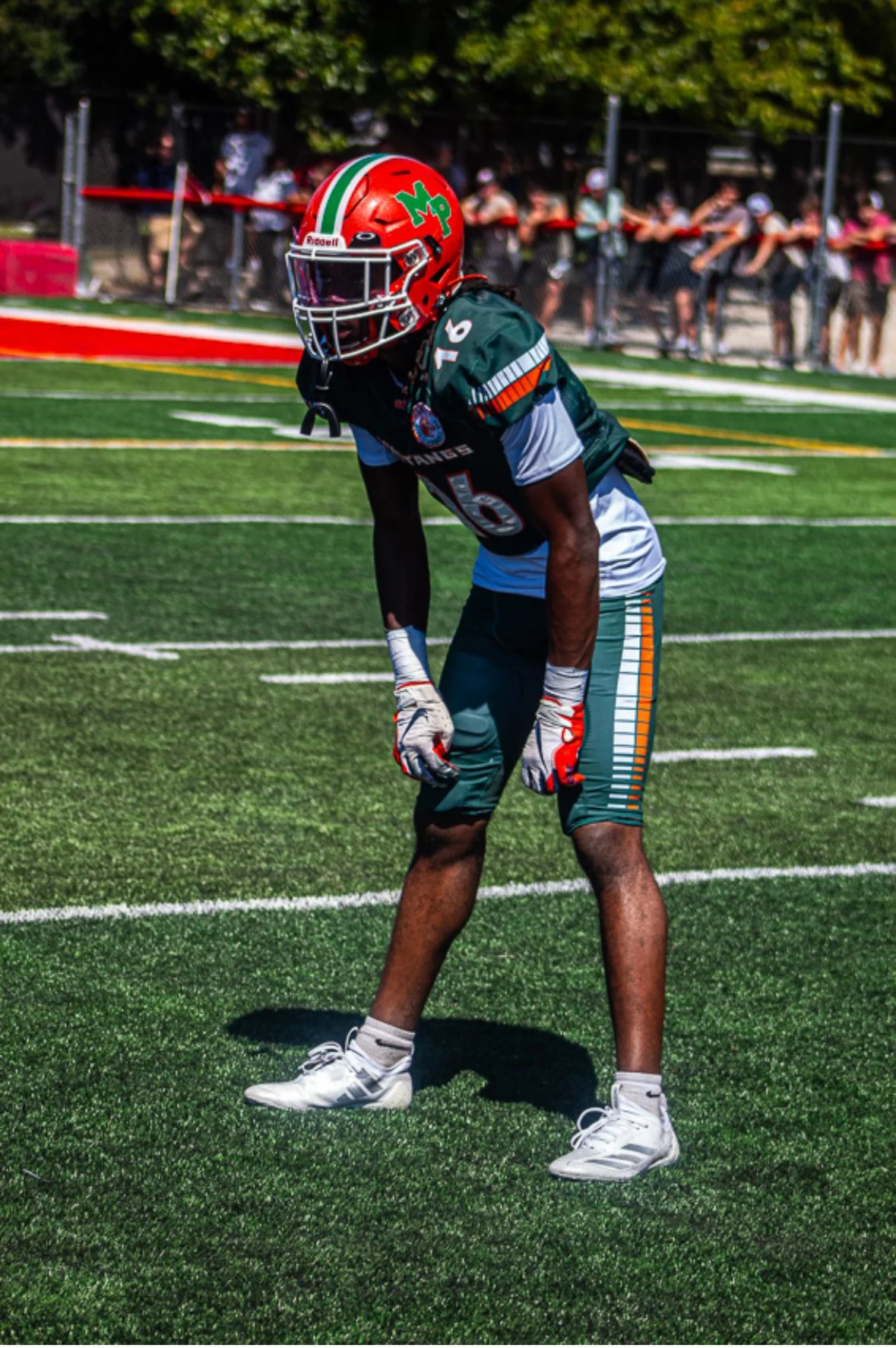 Football player in dark green uniform with orange and white accents, wearing a red helmet with green and white logo, standing on football field during game.