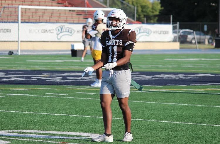 A young football player standing on a football field, wearing a black and white jersey with the word 'SAINTS' and number 11, a helmet, and gloves, with a few other players practicing in the background.