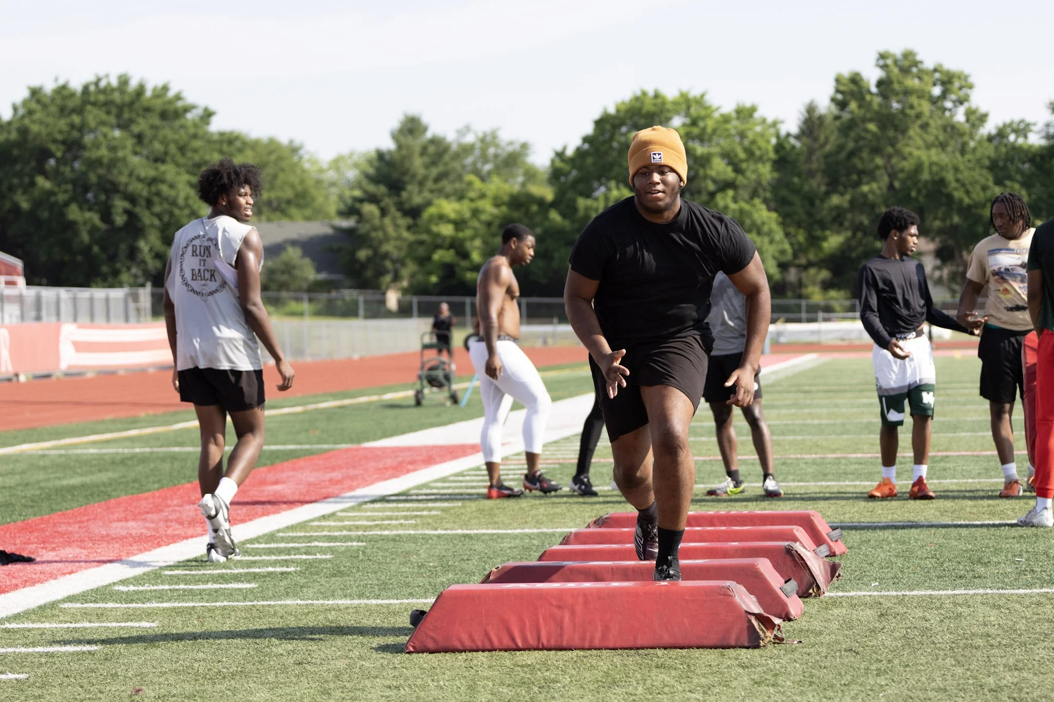 Group of young athletes training on a running track outdoors, with green trees in the background. One athlete in black is jumping over padded hurdles while others stand or walk nearby.