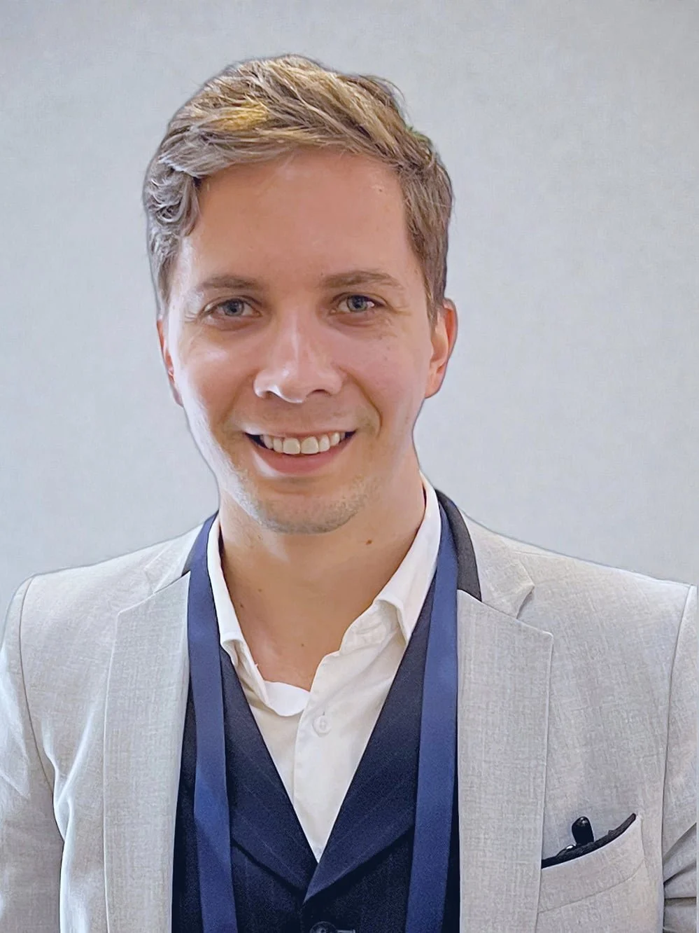 Portrait of a young man with short brown hair, wearing a white dress shirt and navy blazer, against a gray background.
