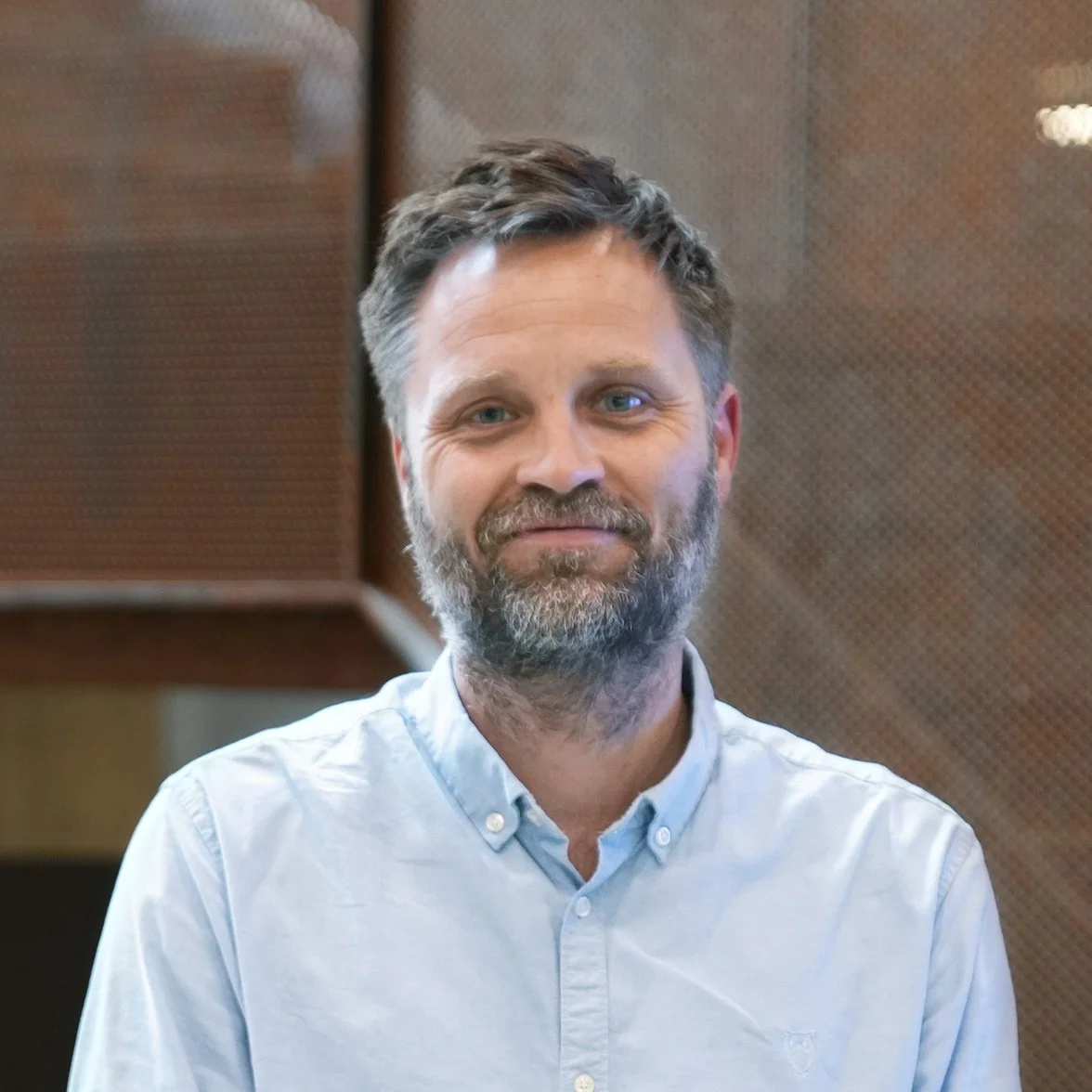 A man with a beard and tousled hair wearing a light blue button-up shirt, standing in front of a wooden and textured wall background.