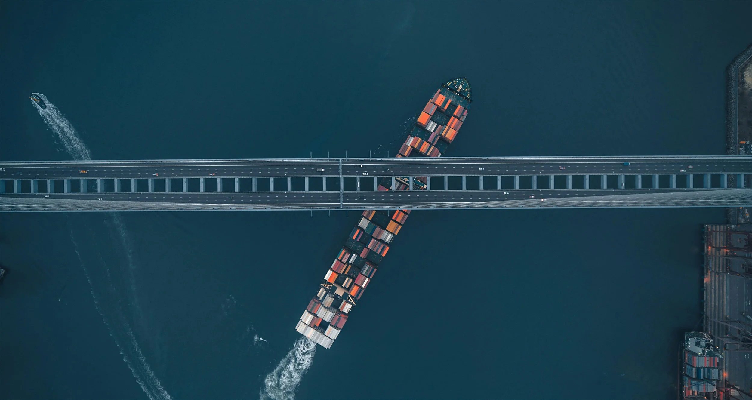 An aerial view of a cargo ship traveling under a bridge with containers stacked on it, while another boat speeds away in the water.