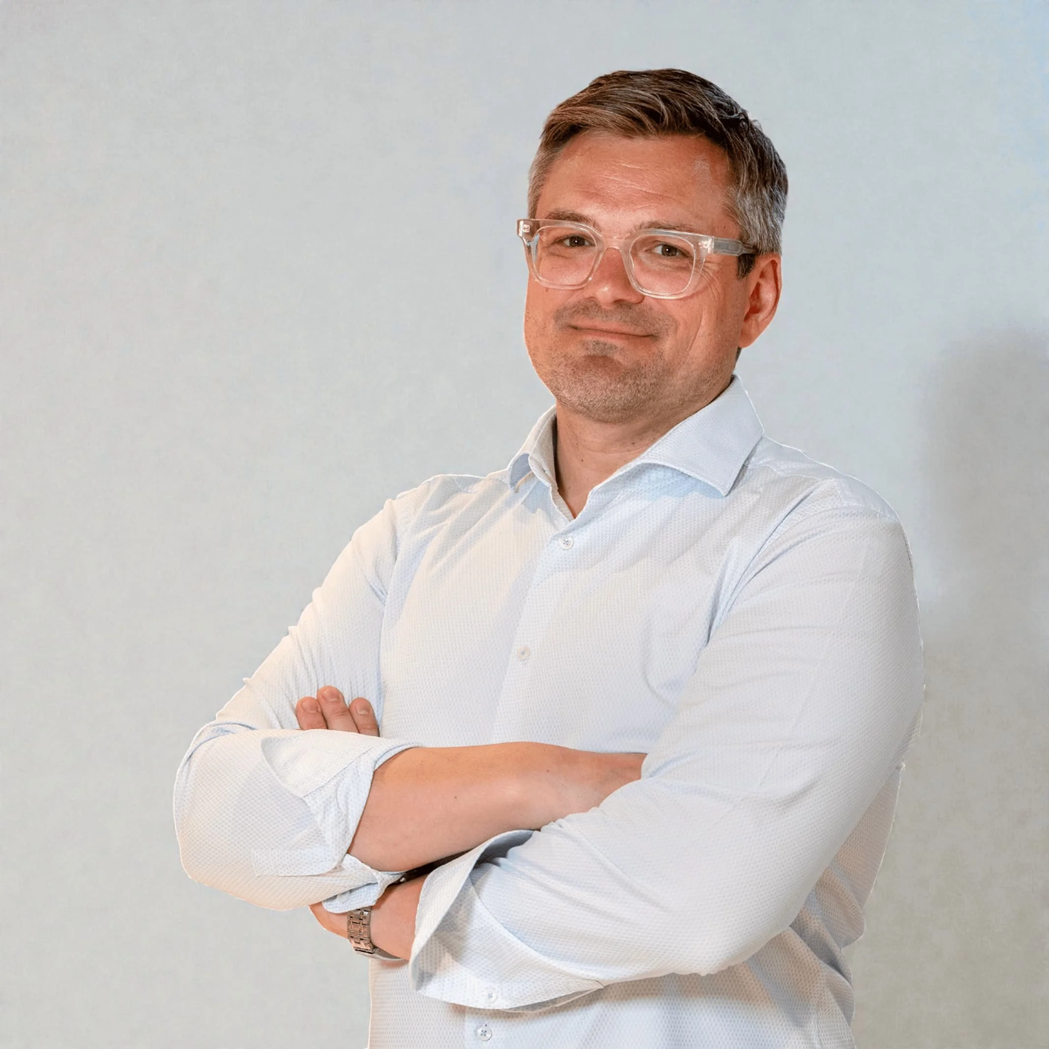 A man wearing glasses and a light shirt stands with arms crossed, smiling slightly, against a plain light background.