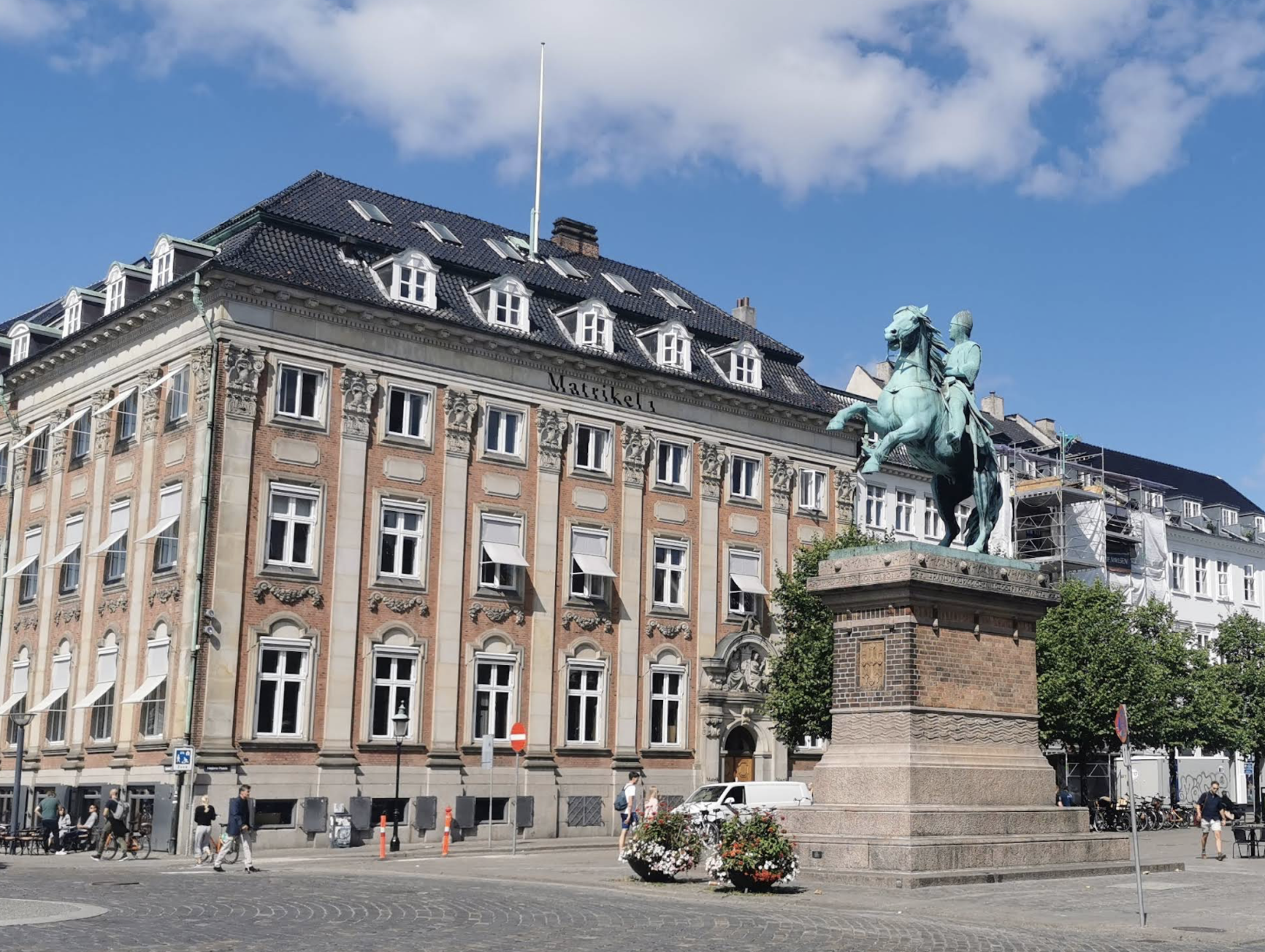A historic building with decorative architecture, a statue of a man on a horse, and a city square with pedestrians.