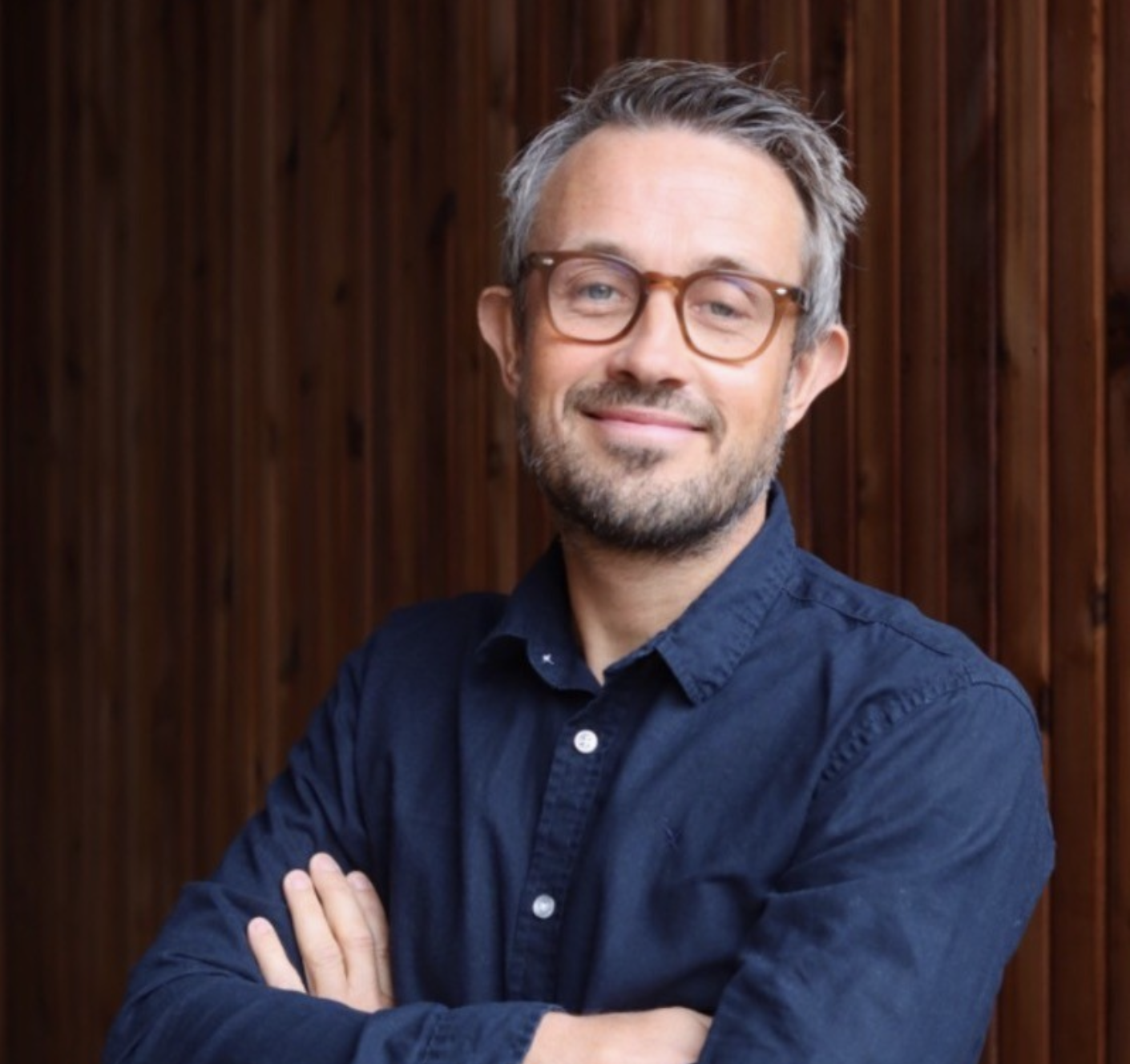 A man with glasses and a beard, smiling with arms crossed, standing in front of a wooden background.