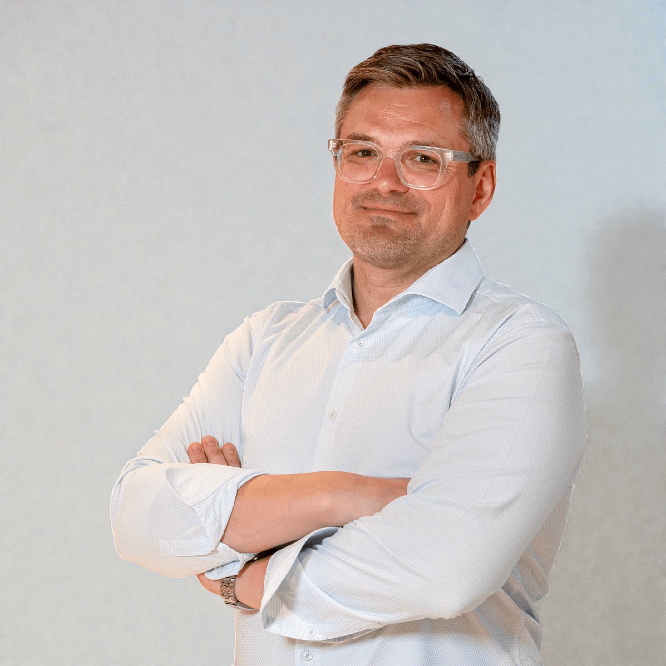 A man wearing glasses and a light shirt stands with arms crossed, smiling slightly, against a plain light background.