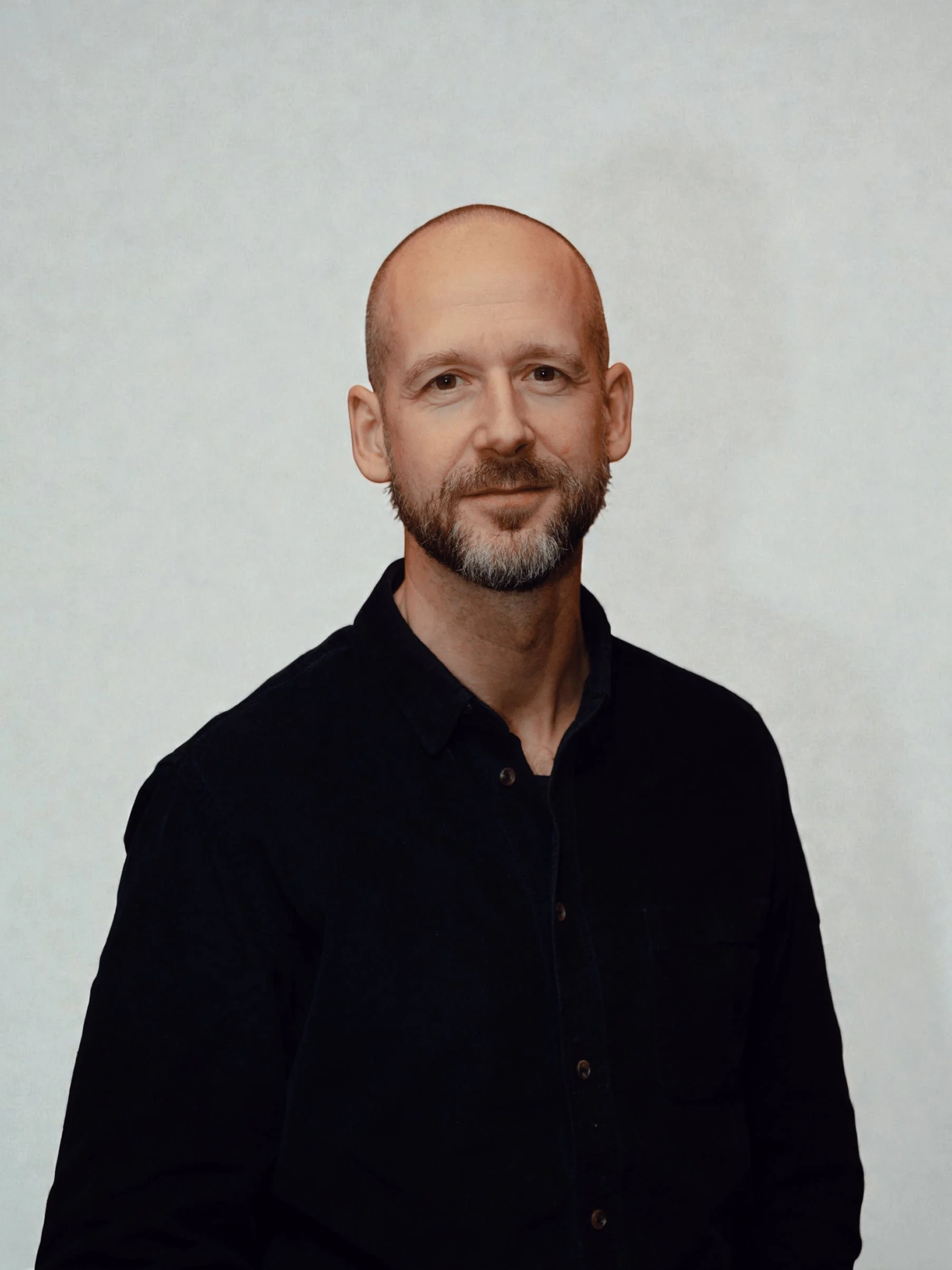 Portrait of a middle-aged man with a shaved head wearing a white collared shirt, smiling softly, against a light blue background.