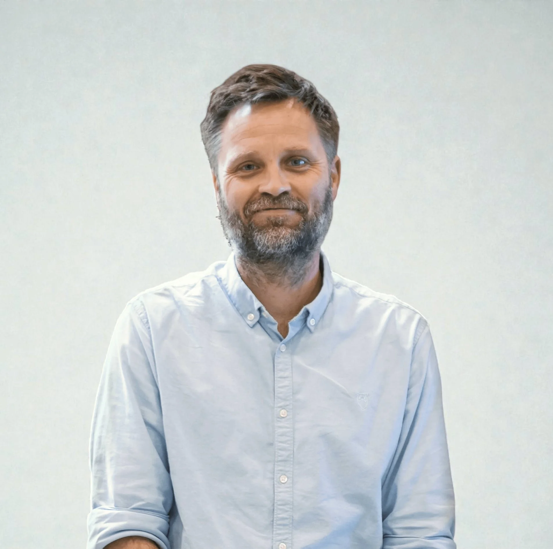 A man with a beard and tousled hair wearing a light blue button-up shirt, standing in front of a wooden and textured wall background.