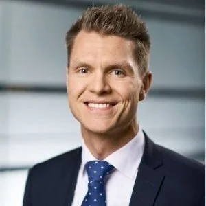 A young man in a suit and tie smiling at the camera, with a blurred office background.