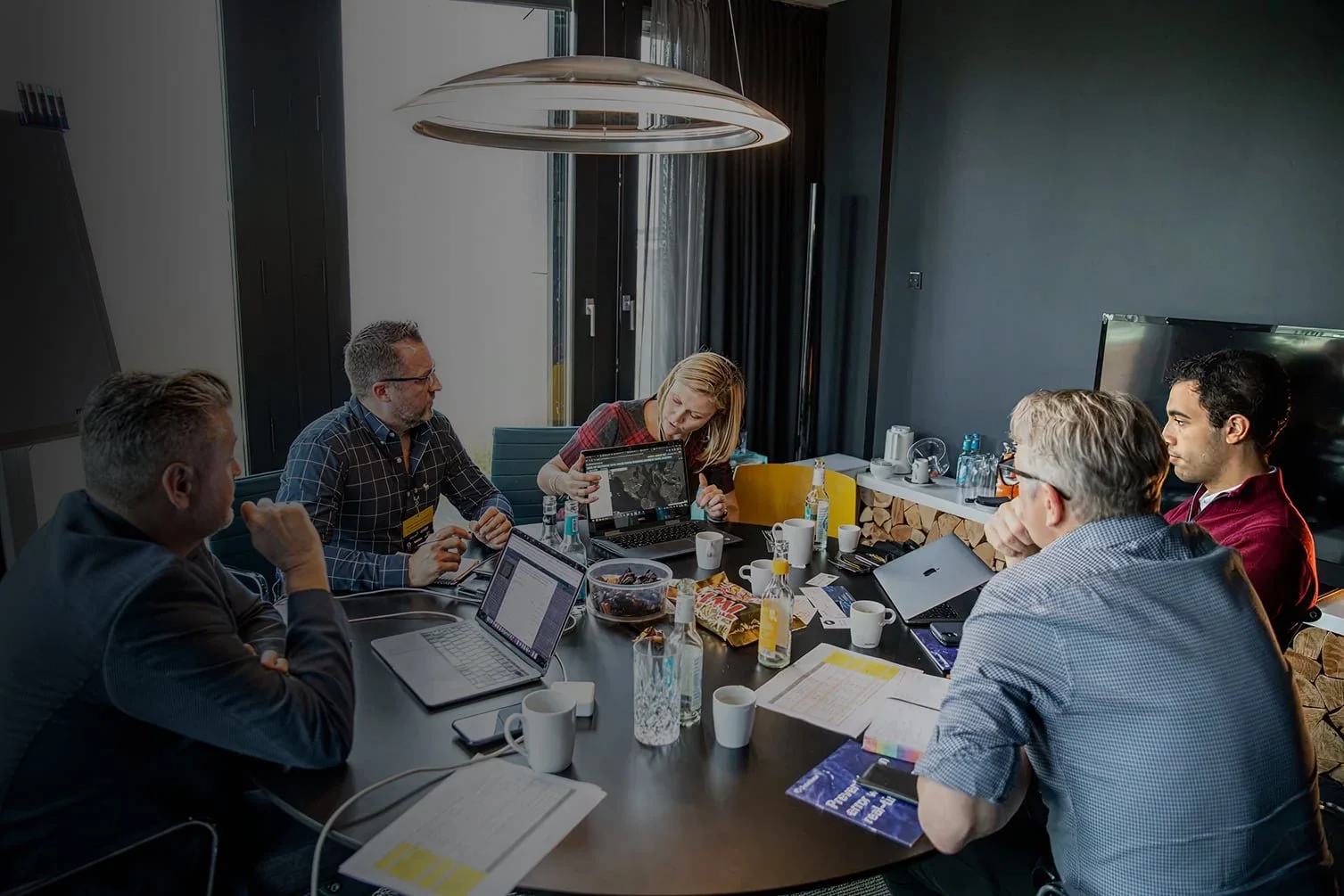 Five people sitting around a conference table in a modern office, engaged in a discussion. One woman is showing a presentation on a laptop. The table has notebooks, laptops, bottles, and cups. Large window with blinds in the background.