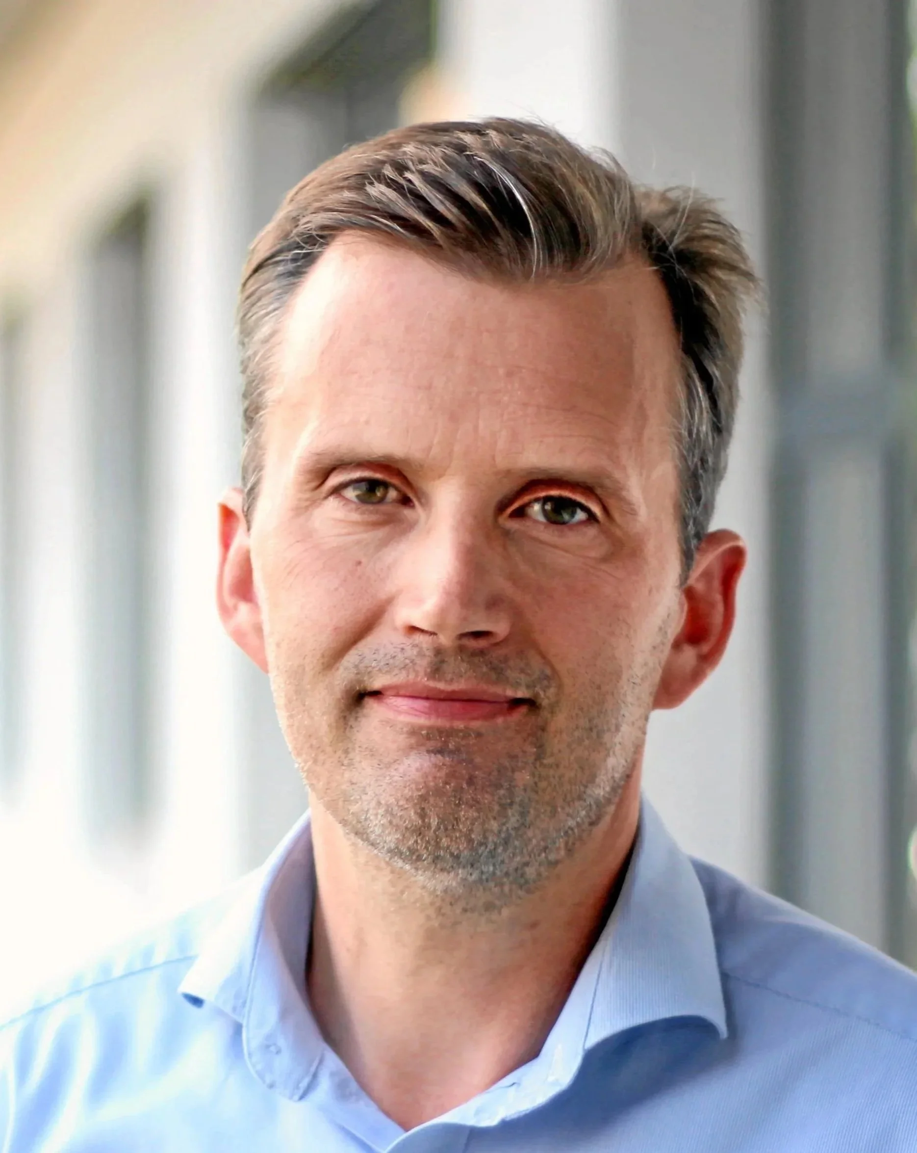 Close-up of a middle-aged man with short brown hair, light stubble, wearing a light blue collared shirt, standing in front of blurred windows.