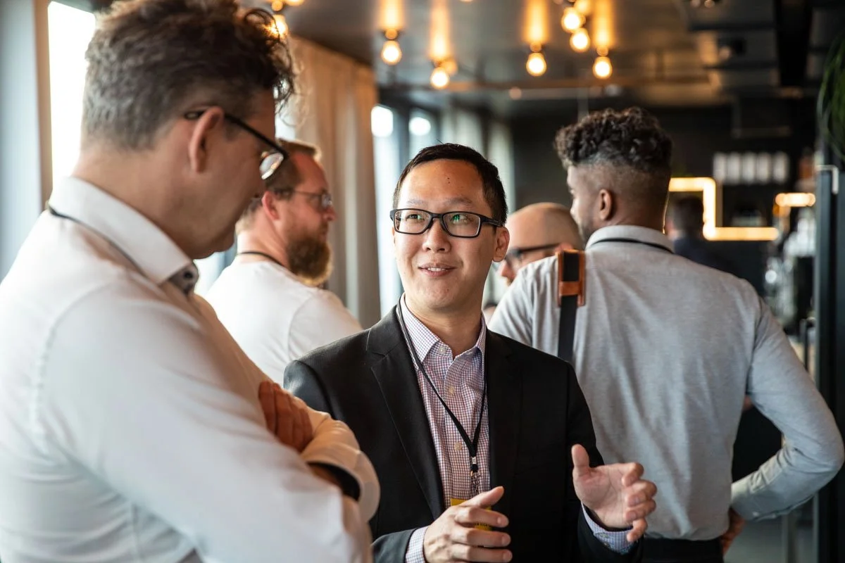 Group of professionals networking and conversing at an indoor event, with warm lighting and modern decor in the background.