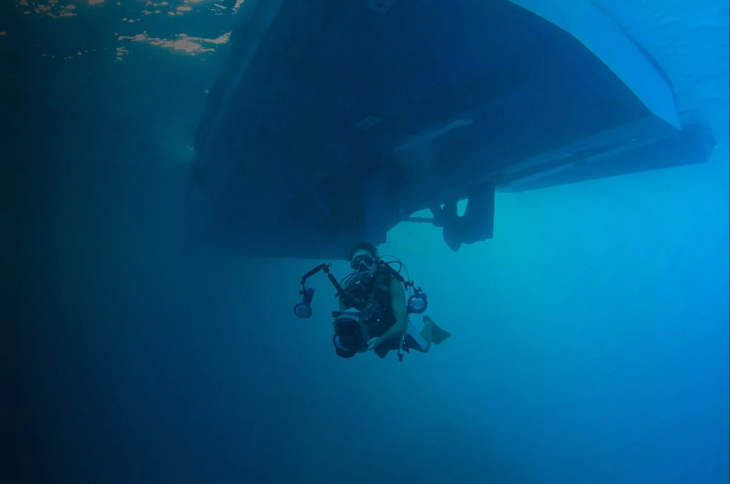Underwater photo of a scuba diver with a camera, swimming beneath a boat in the ocean.