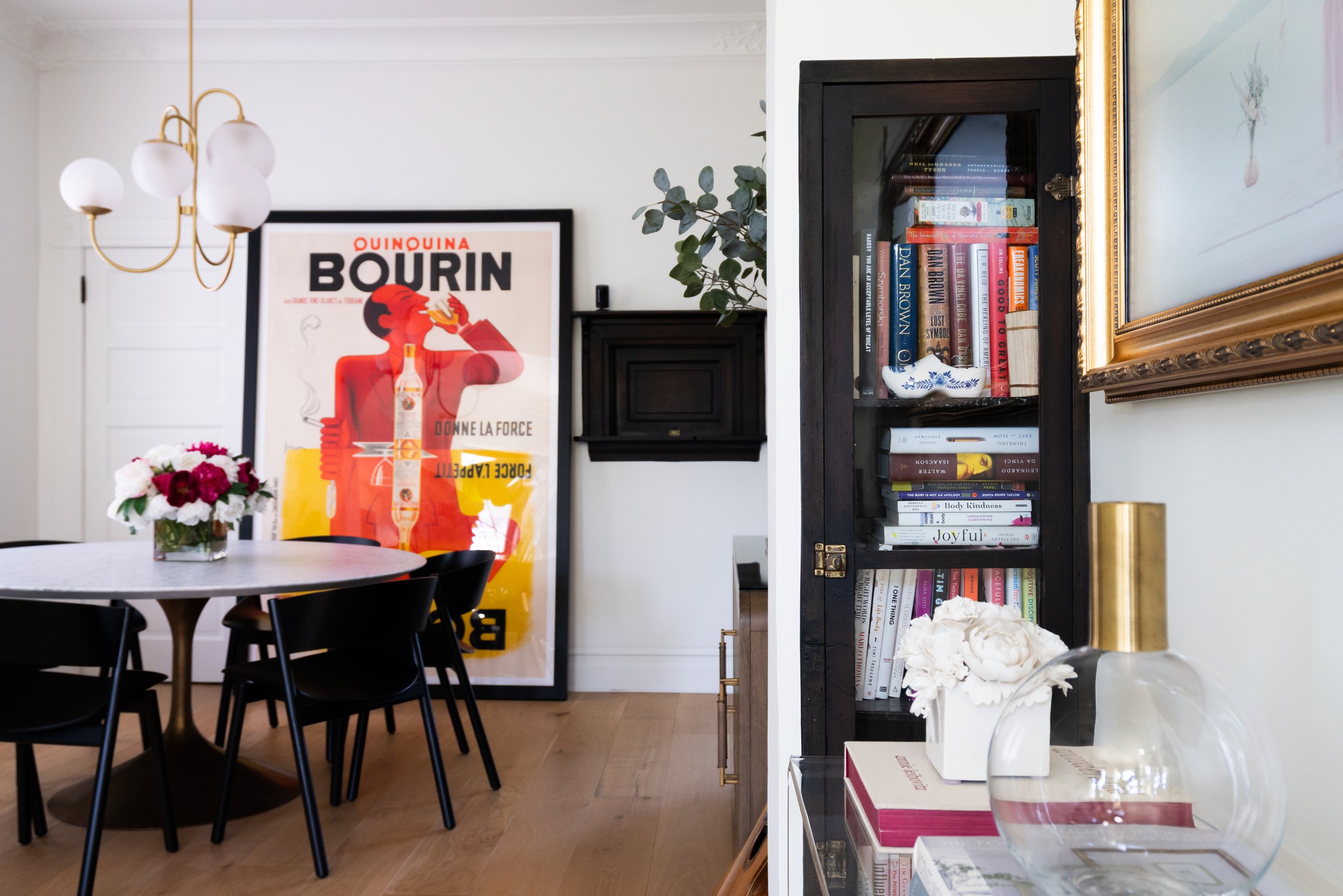 interior design of a dining room with a marble dining table, black wood chairs, large piece of art against the wall, floral on the table below a grand chandelier