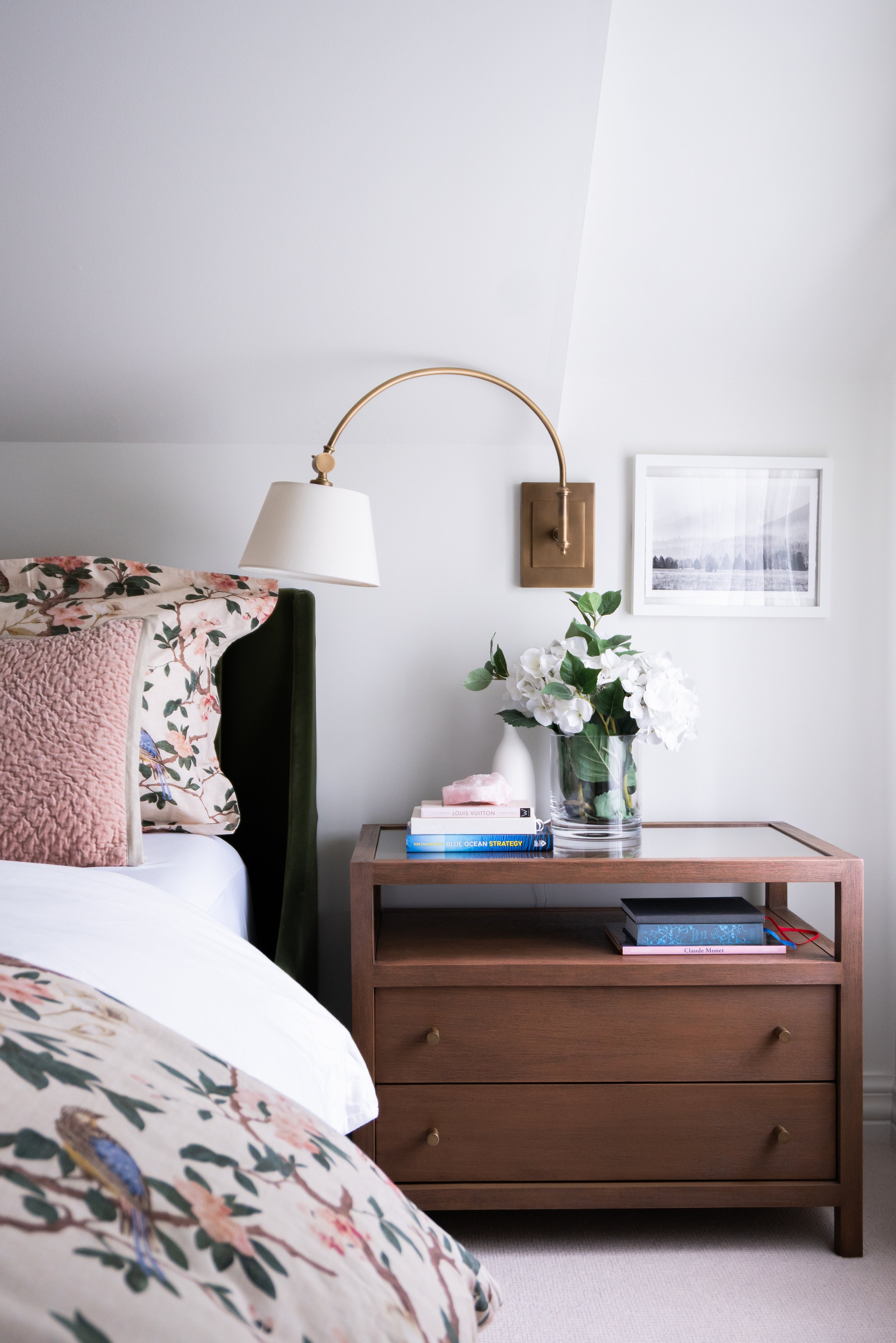 interior design of a light and airy bedroom with a green headboard, sconce on the wall and walnut end table
