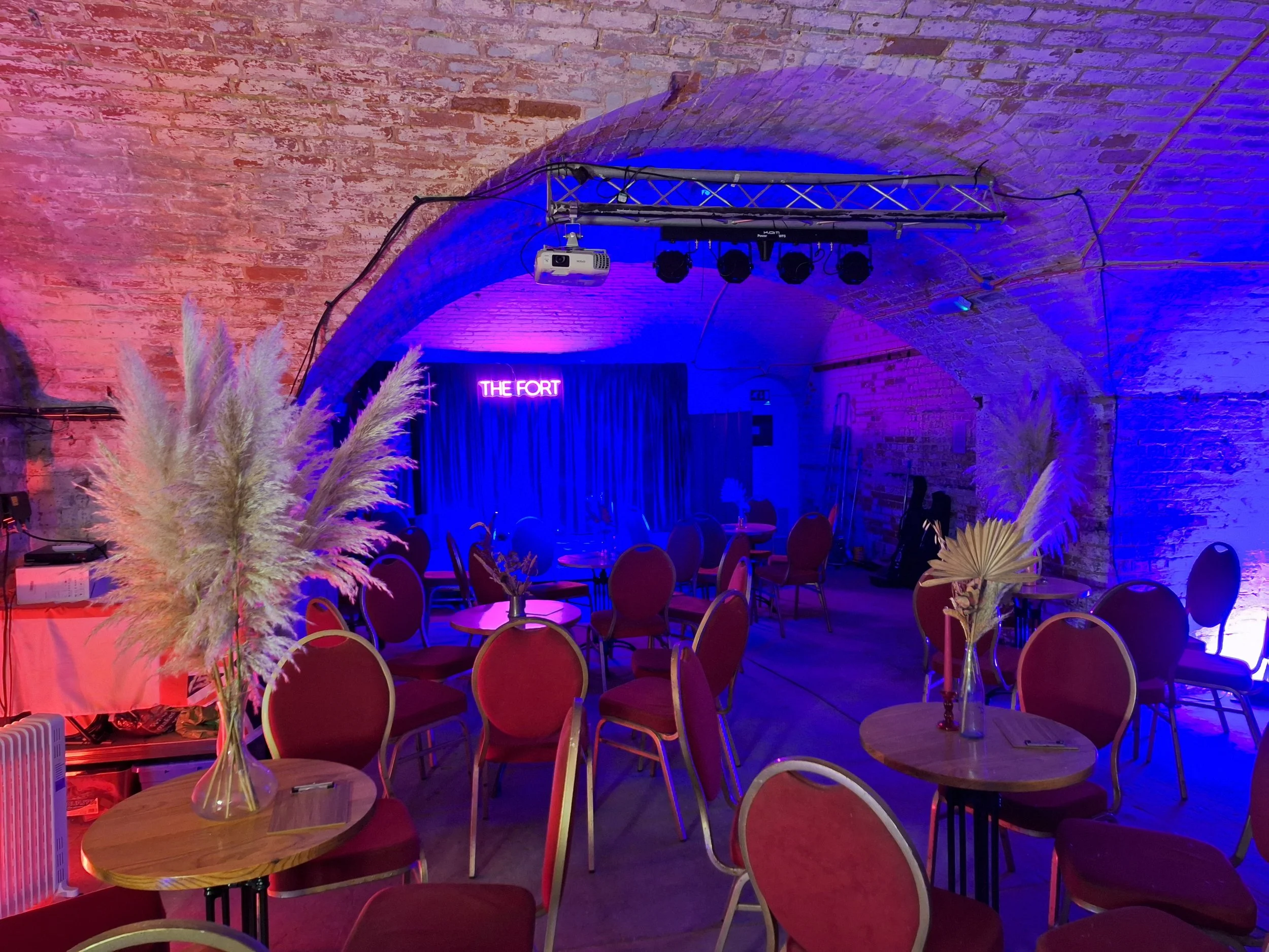 Event space with tables and chairs, decorated with tall blurred pampas grass in vases, illuminated with pink and blue lighting, a neon sign reading 'THE FORT' on the stage, brick arched ceiling, projector hanging from the ceiling, and stage area with dark curtains.