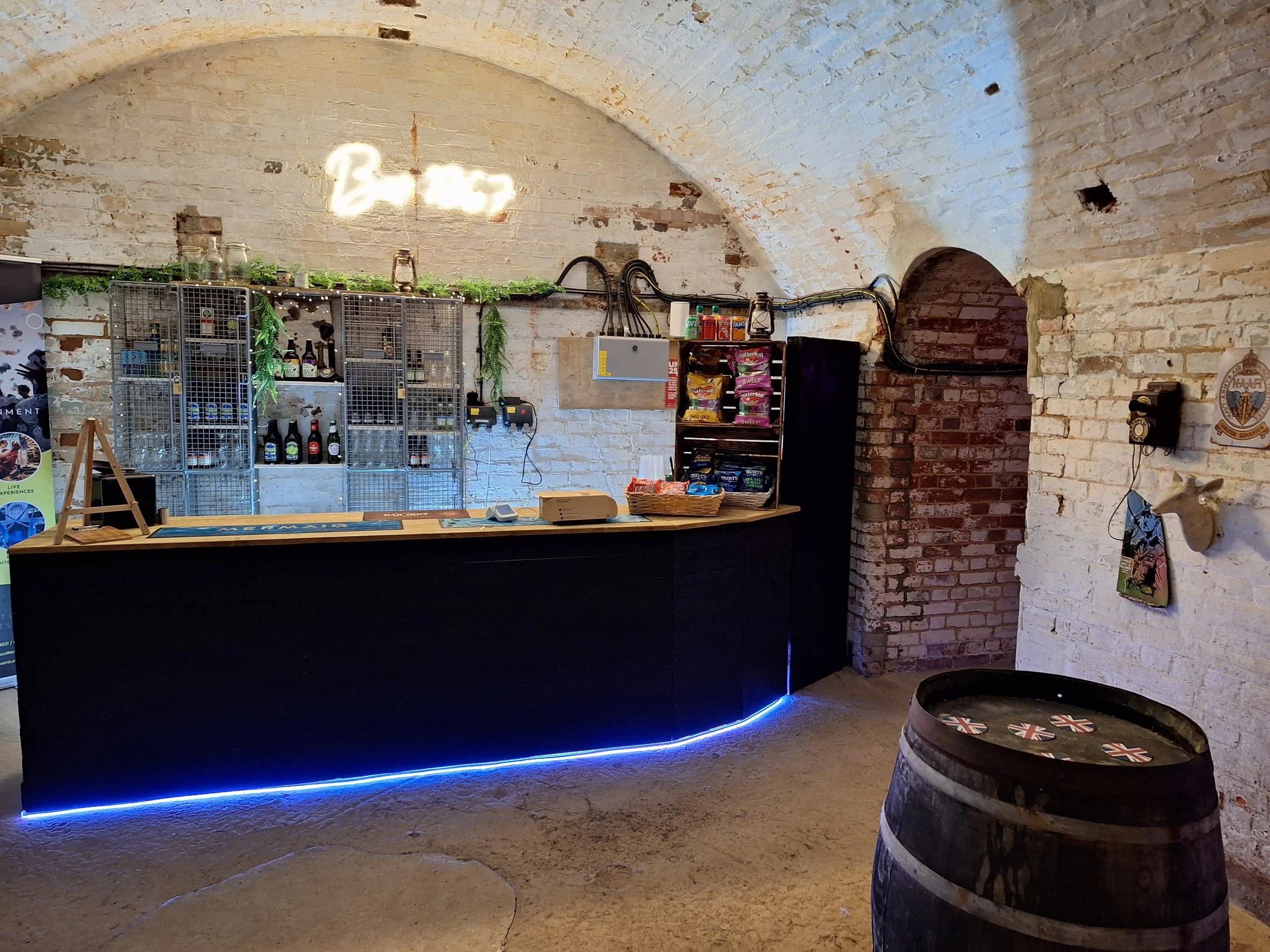 Interior of a rustic brick building with a bar counter. There is a neon sign that reads 'Beer' on the wall above the bar. Shelves behind the counter contain bottles, jars, and snacks. A barrel with UK flags is in the foreground, and a small platform with blue LED lighting runs along the bottom edge of the bar.